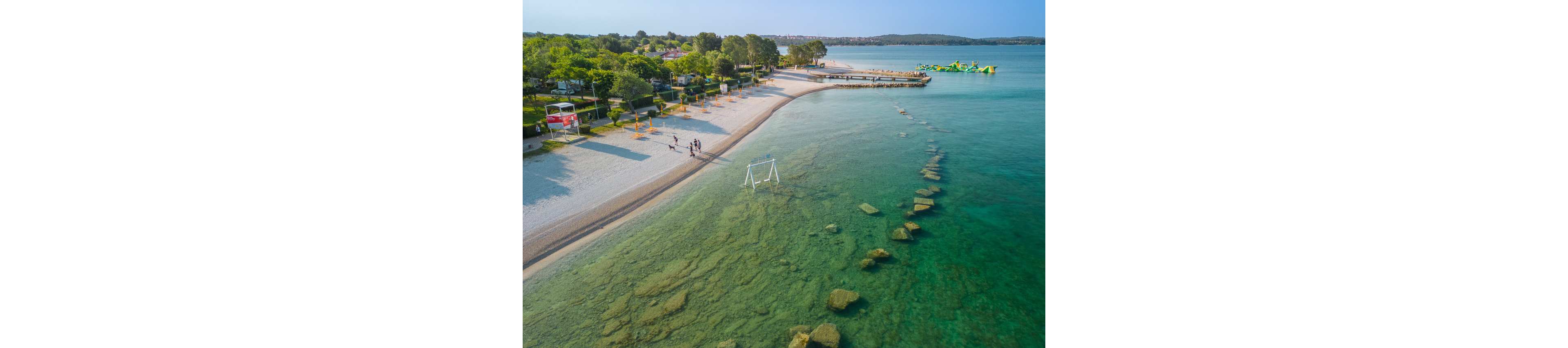 An aerial view captures a tranquil beach with clear turquoise water and a sandy shoreline. A small group of people walks along the beach near shaded seating areas and trees. In the shallow water, a white goal-like frame stands among visible rocks, adding a playful element. Farther out, an inflatable water park floats on the calm sea, while a distant coastline and low hills frame the horizon.