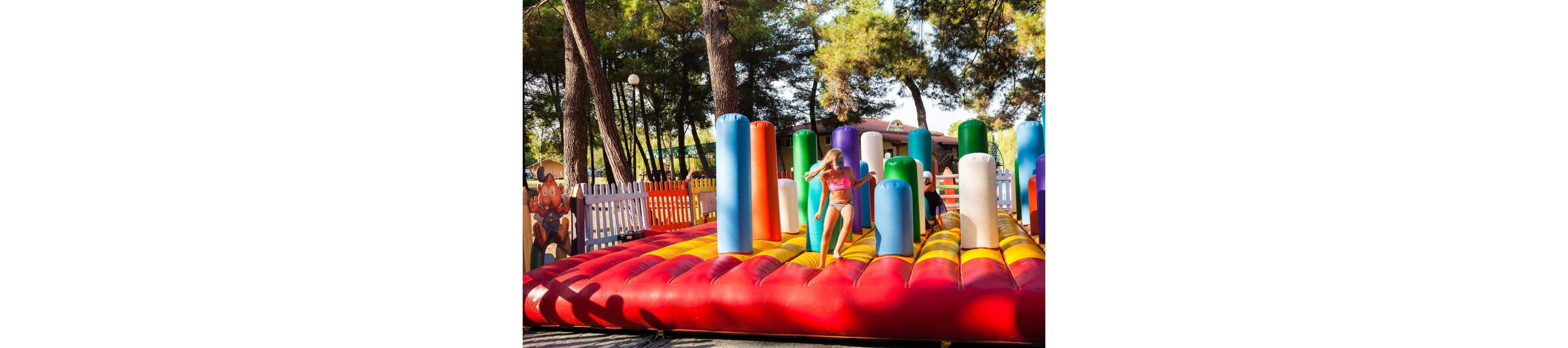 A young girl in a swimsuit plays on a bright inflatable structure with tall, multicolored pillars. Another child is partially visible behind the columns, also enjoying the attraction. The playground is set outdoors among tall trees, with white picket fencing and cartoon decorations in the background. Warm sunlight and vivid colors create a cheerful, summery atmosphere.