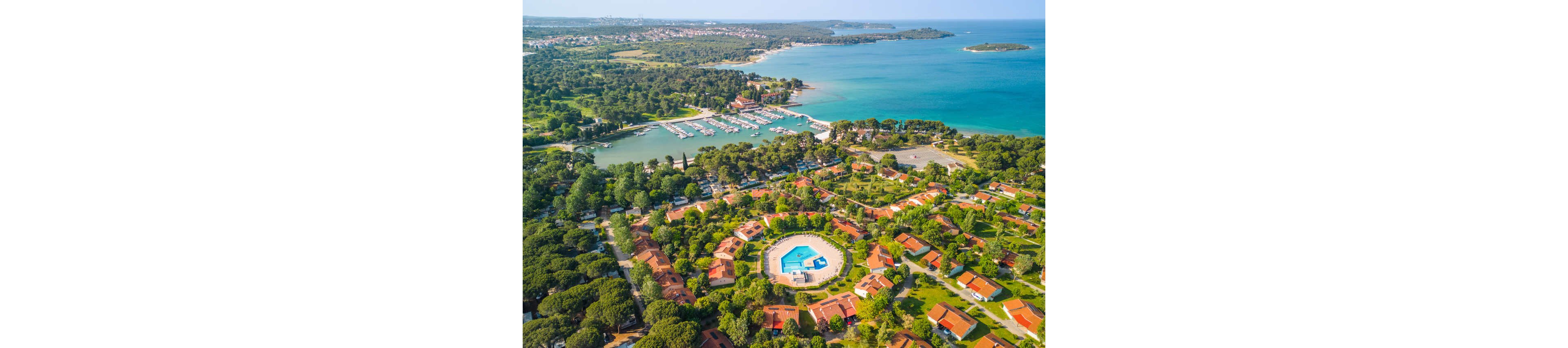 A large coastal holiday resort is captured from above, showing a central hexagonal swimming pool surrounded by villas and dense green trees. The complex sits next to a marina filled with boats and yachts, with calm turquoise sea stretching into the distance. Red-roofed houses and winding roads are visible among the greenery, suggesting a peaceful vacation community. The overall scene conveys a sunny, relaxed Mediterranean or Adriatic seaside atmosphere.