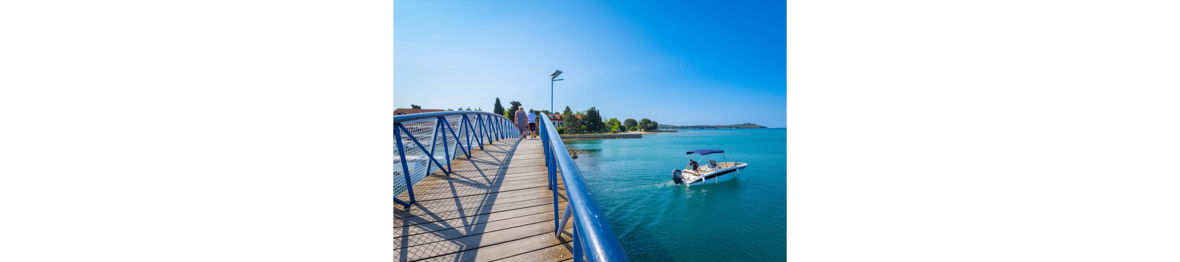 A small motorboat cruises through calm turquoise water near a coastal town. On the left, people walk along a blue-railed pedestrian bridge that extends over the bay. Bright sunlight and a clear blue sky create a vivid, summery atmosphere. Trees and low buildings line the distant shoreline, adding to the relaxed seaside setting.