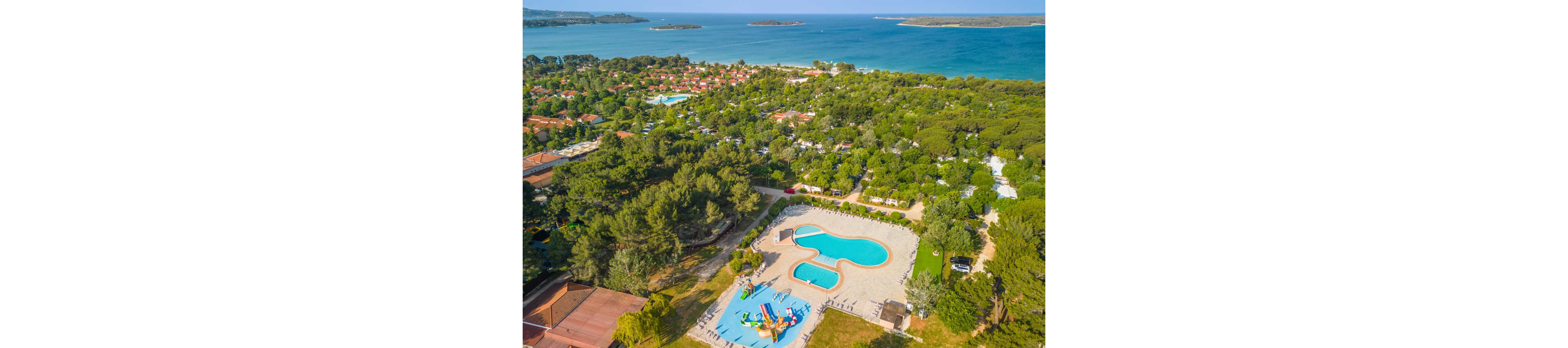 An aerial view shows a large coastal holiday resort surrounded by dense green trees. In the foreground, there is a bright blue swimming pool complex with multiple irregularly shaped pools and a colorful water play area. Small buildings, campsites, and pathways are scattered among the trees, leading toward the shoreline. In the background, the turquoise sea and several small islands create a scenic coastal landscape under clear skies.