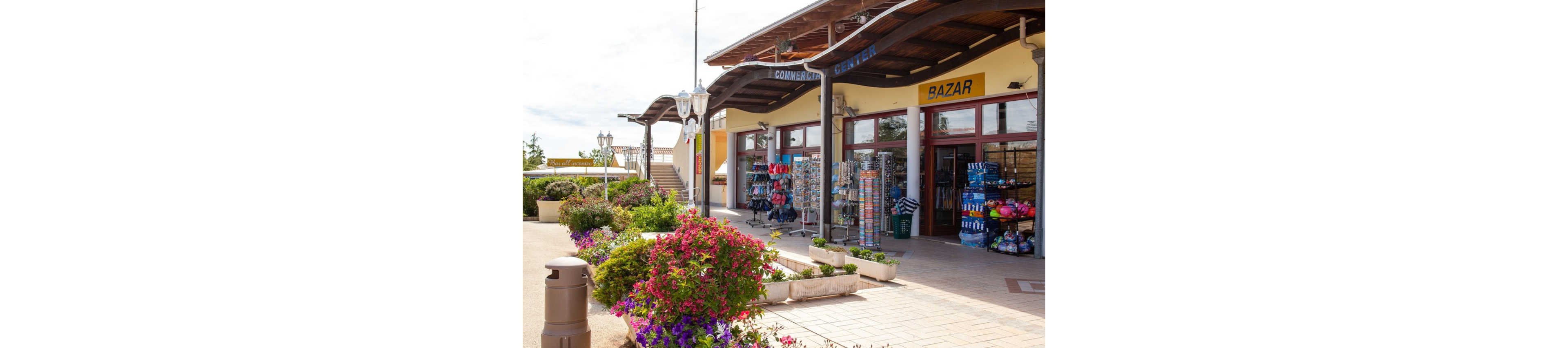 An outdoor shopping area features a bazaar and commercial center with open storefronts. Colorful racks of beach toys, towels, and souvenirs line the entrances. Flowering plants in large planters decorate the walkway, adding vibrant pink and purple blooms. The scene is bright and sunny, suggesting a relaxed vacation or resort atmosphere.
