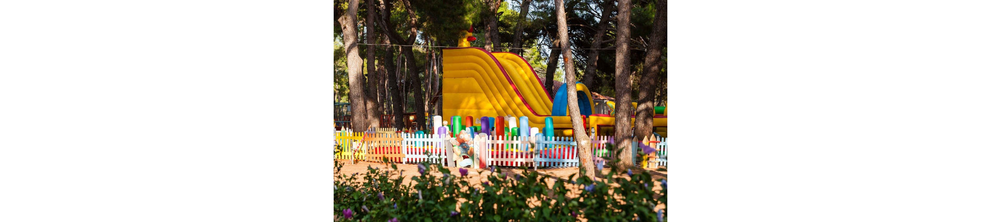 A large yellow inflatable slide stands in the middle of a fenced playground surrounded by tall pine trees. Brightly colored plastic posts and a white picket fence enclose the play area. A small child is visible near the fence, adding a sense of scale and activity. The scene is sunlit, with vivid primary colors contrasting against the natural green forest background.
