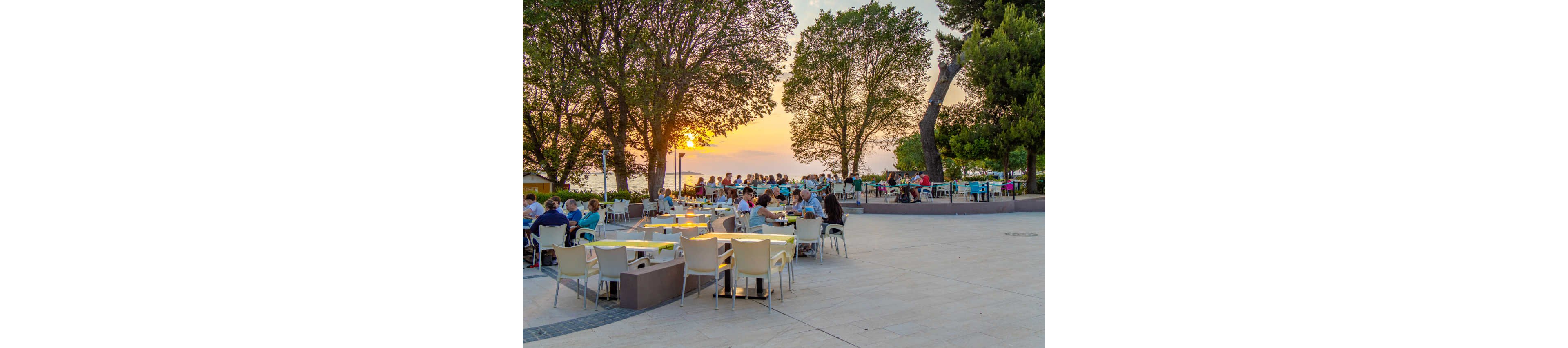 People sit at an outdoor café terrace near the sea as the sun sets behind tall trees. Groups of diners relax at white tables and chairs, enjoying food and drinks in the warm evening light. The scene overlooks a waterfront promenade with more people walking and socializing in the background. Soft golden sunlight and green foliage create a calm, vacation-like atmosphere.