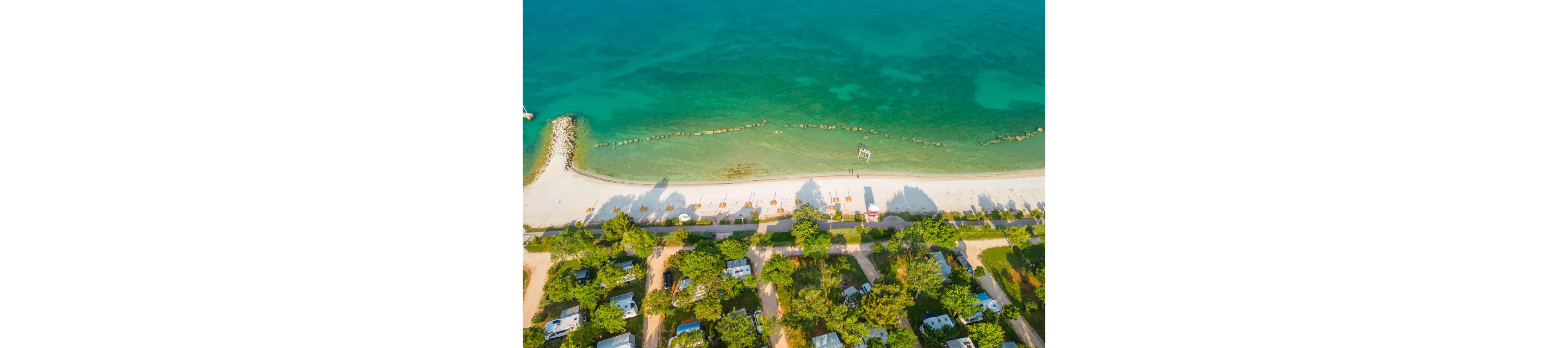 An aerial view shows a beachside campground next to a clear turquoise sea. A row of sun loungers and umbrellas lines the sandy shoreline, casting long shadows toward the water. Small cabins, RVs, and tents are arranged among green trees behind the beach. The calm water, bright sand, and organized camping area create a relaxed summer holiday atmosphere.