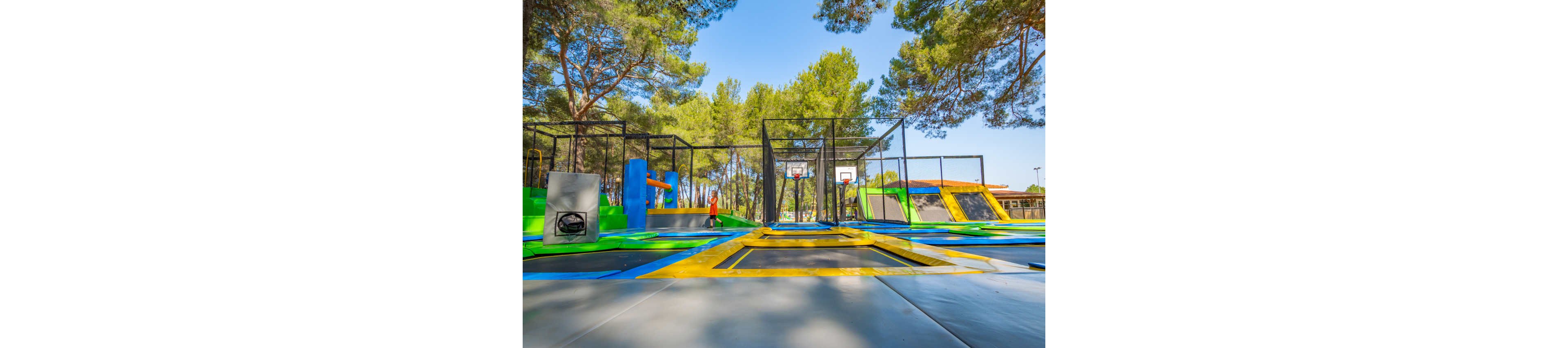 An outdoor trampoline park features multiple colorful trampolines arranged in rows. A child stands near the center, surrounded by safety nets and basketball hoops integrated into the play area. Tall green trees and clear blue sky create a bright, sunny atmosphere. The scene conveys active fun, recreation, and family-friendly entertainment in a natural setting.