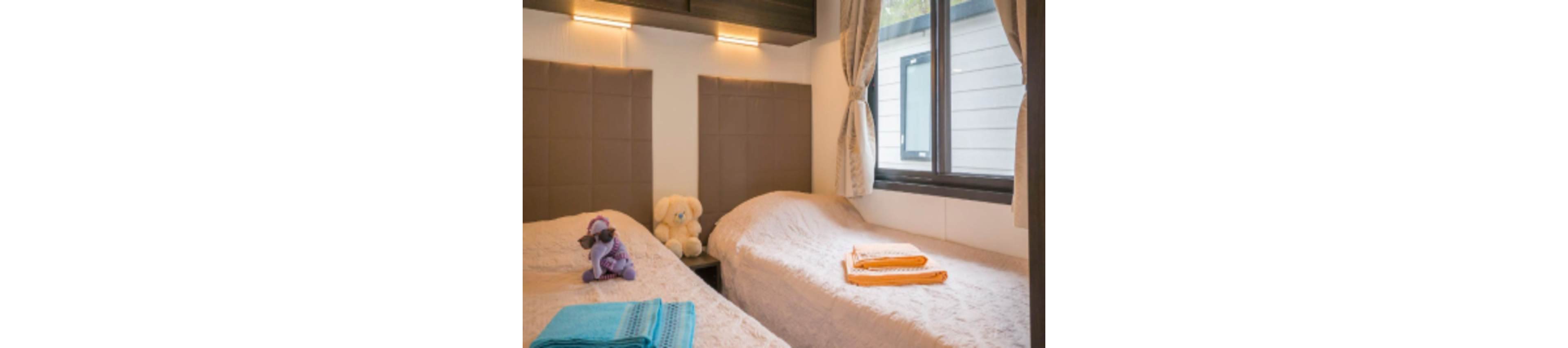 A small, cozy bedroom features two single beds with light-colored bedspreads. Soft toys, including a teddy bear and a plush figure, sit on one of the beds, suggesting a child-friendly space. Folded towels in bright colors are neatly placed at the foot of each bed. Warm wall lights, padded headboards, and a large window with curtains create a comfortable and inviting atmosphere.