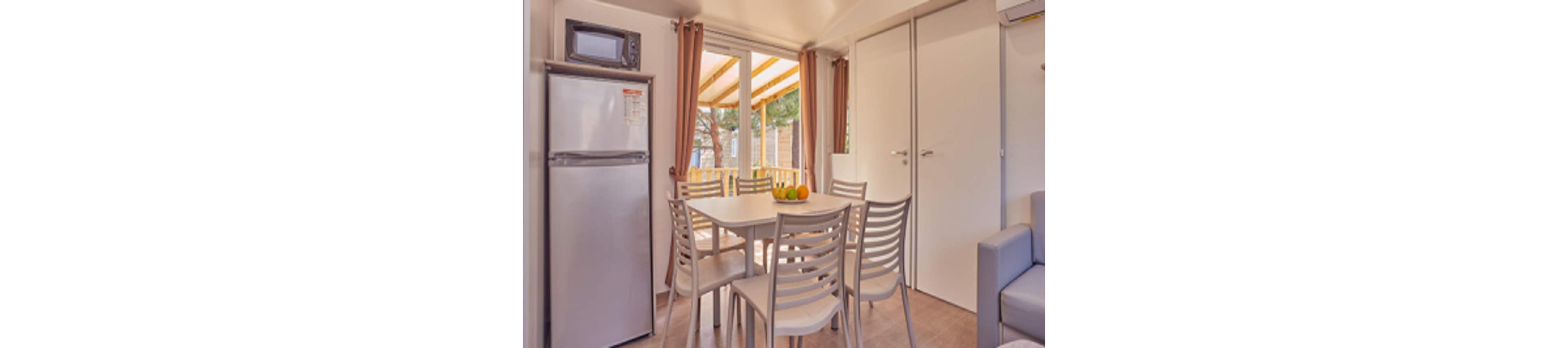A small dining area with a square table and four chairs is set inside a compact kitchen space. A tall refrigerator with a microwave on top stands to the left, while a light-colored sofa is partially visible on the right. Large glass doors with curtains open to a covered terrace, allowing warm natural light to fill the room. A bowl of fruit on the table adds a touch of color to the neutral interior.
