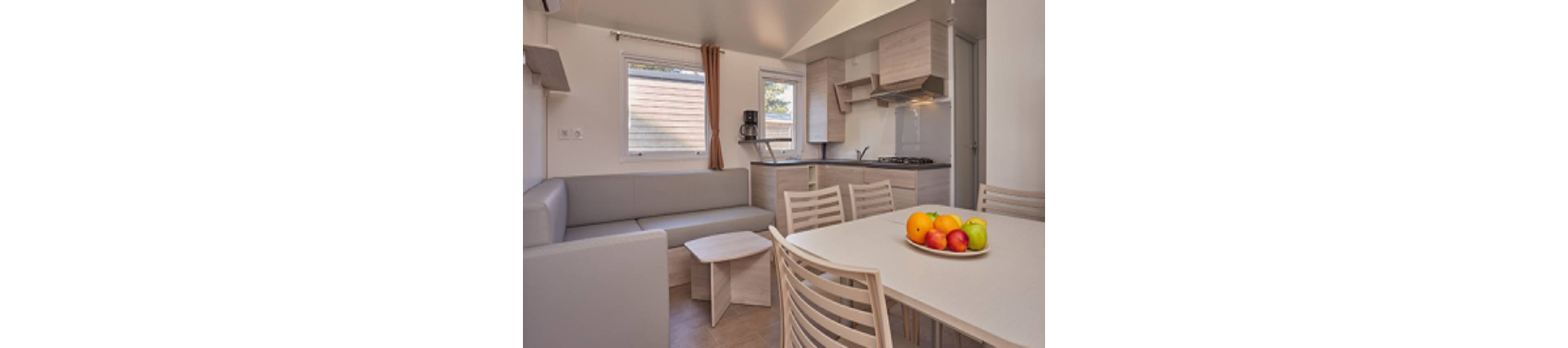 A compact modern kitchen and dining area is shown inside a small home or vacation cabin. Light wood cabinets, a built-in stove, and a corner sink create a clean, functional cooking space. A gray L-shaped sofa sits beside a small coffee table near two windows with simple curtains. In the foreground, a dining table with matching chairs holds a bowl of colorful fruit, adding a bright accent to the neutral decor.