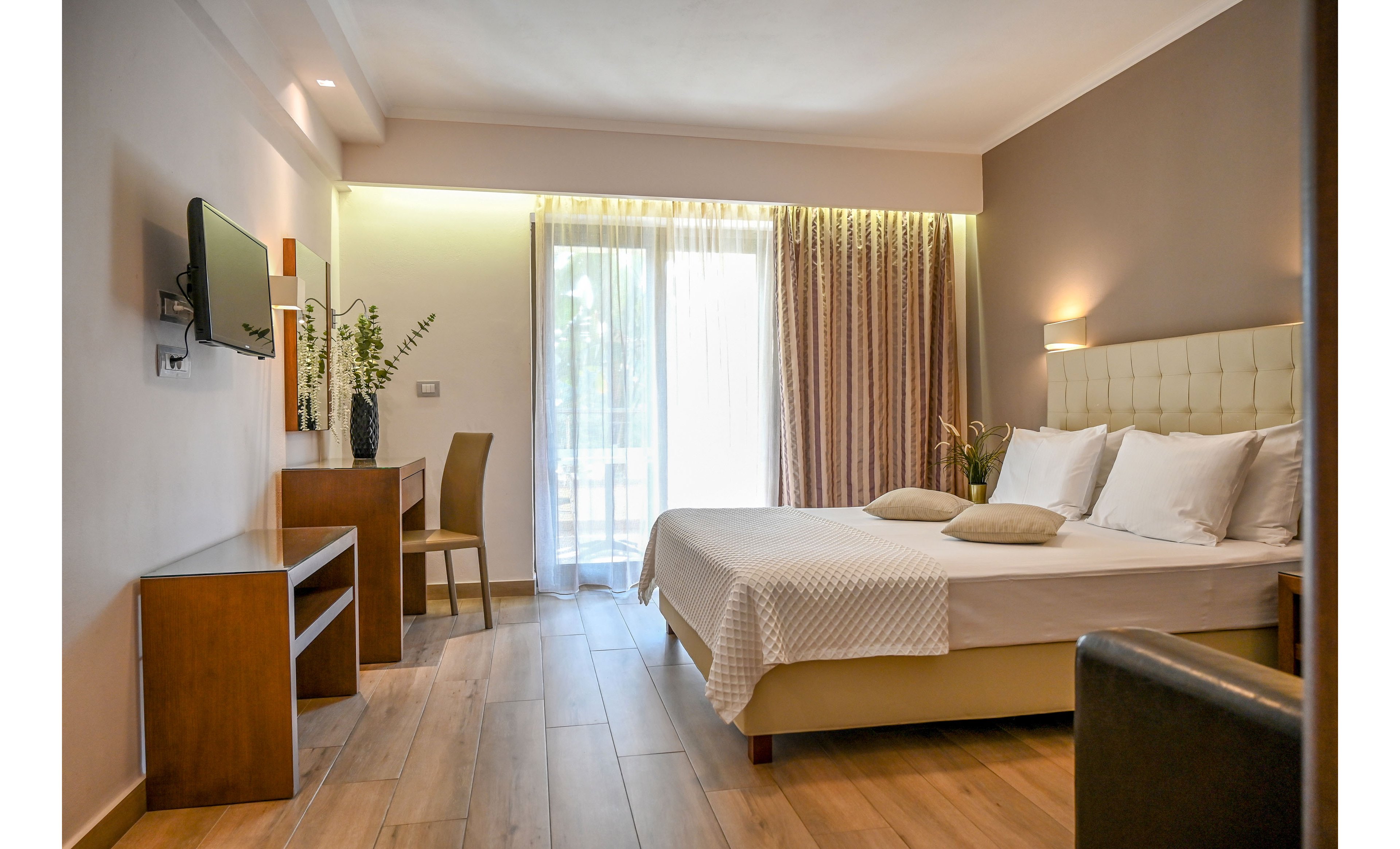 A contemporary hotel-style bedroom features a double bed with white linens and beige cushions. The room includes a wooden desk with a chair, decorative plants, and a wall-mounted television. Floor-to-ceiling glass doors lead to a balcony, allowing natural light to fill the space through sheer curtains. Warm wood flooring and neutral wall tones create a calm, inviting atmosphere.