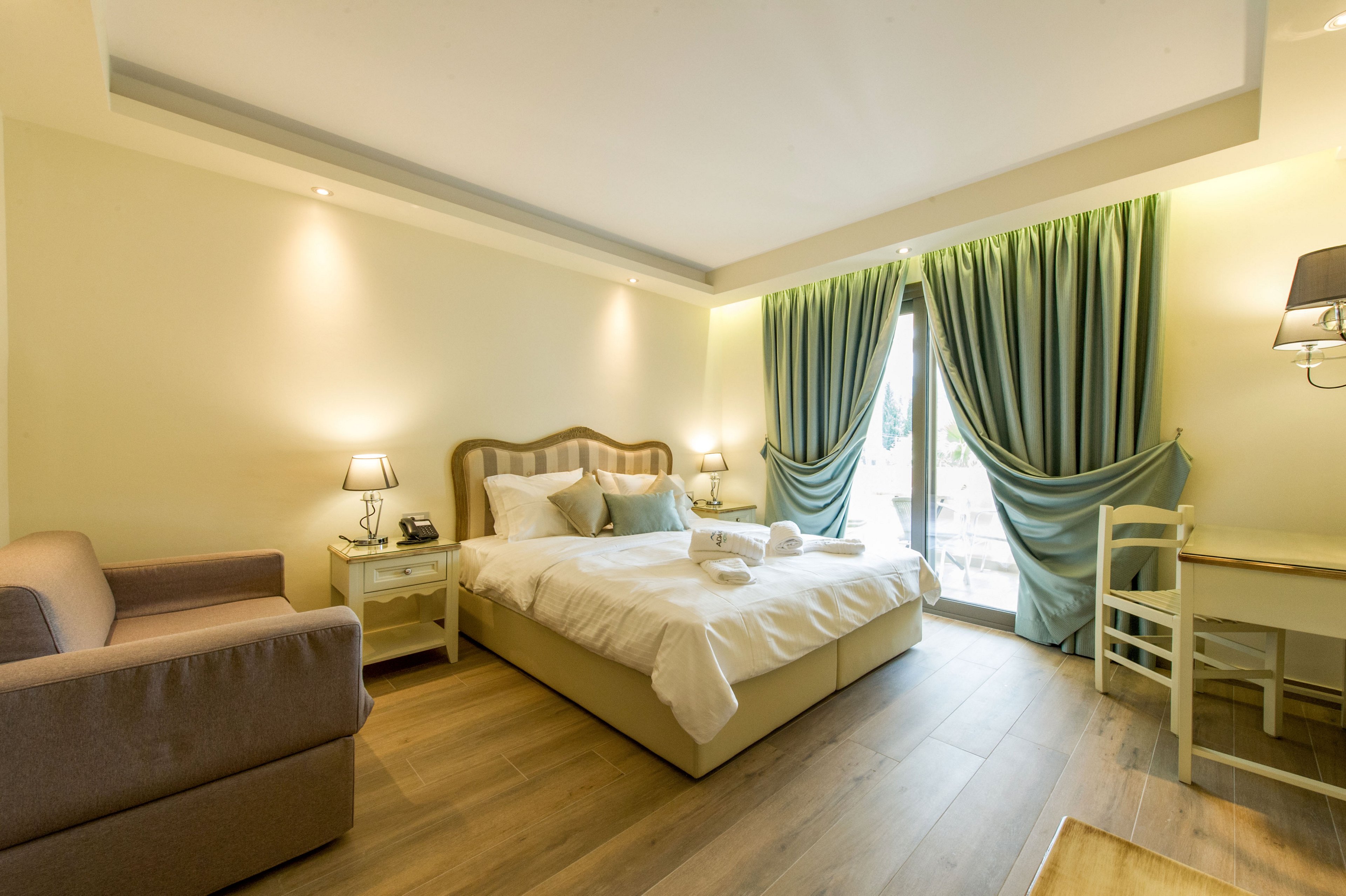 A neatly made double bed sits in a bright, modern hotel room with soft neutral walls and wooden flooring. Two bedside tables with lamps flank the bed, and folded towels are placed on top of the duvet. A sofa and a simple desk with a chair complete the comfortable interior. Large glass doors with green curtains open to a balcony, allowing natural light to fill the space and create an inviting atmosphere.