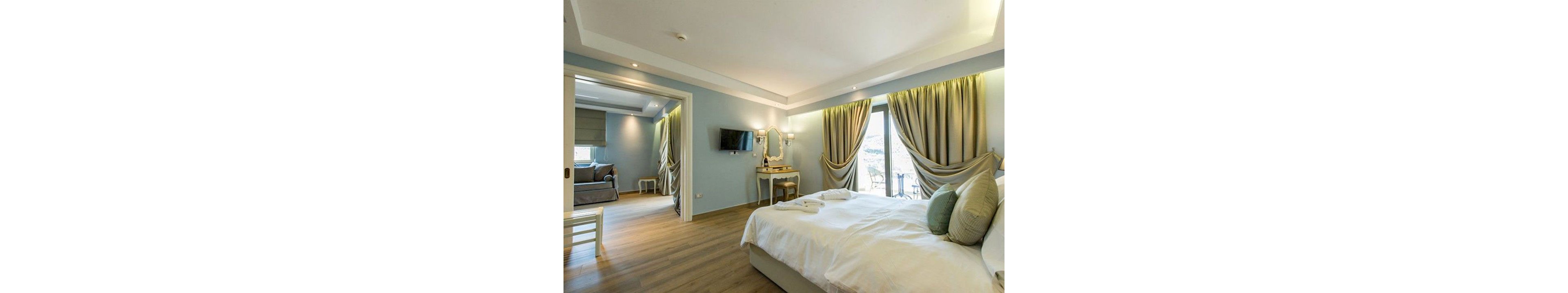 A spacious hotel bedroom features a neatly made bed with white linens and soft green accent pillows. Large windows with flowing curtains open to a balcony, allowing natural light to fill the room. A wall-mounted television and a vanity table with a mirror sit opposite the bed. An adjoining sitting area with additional seating is visible through an open doorway, creating a comfortable suite layout.