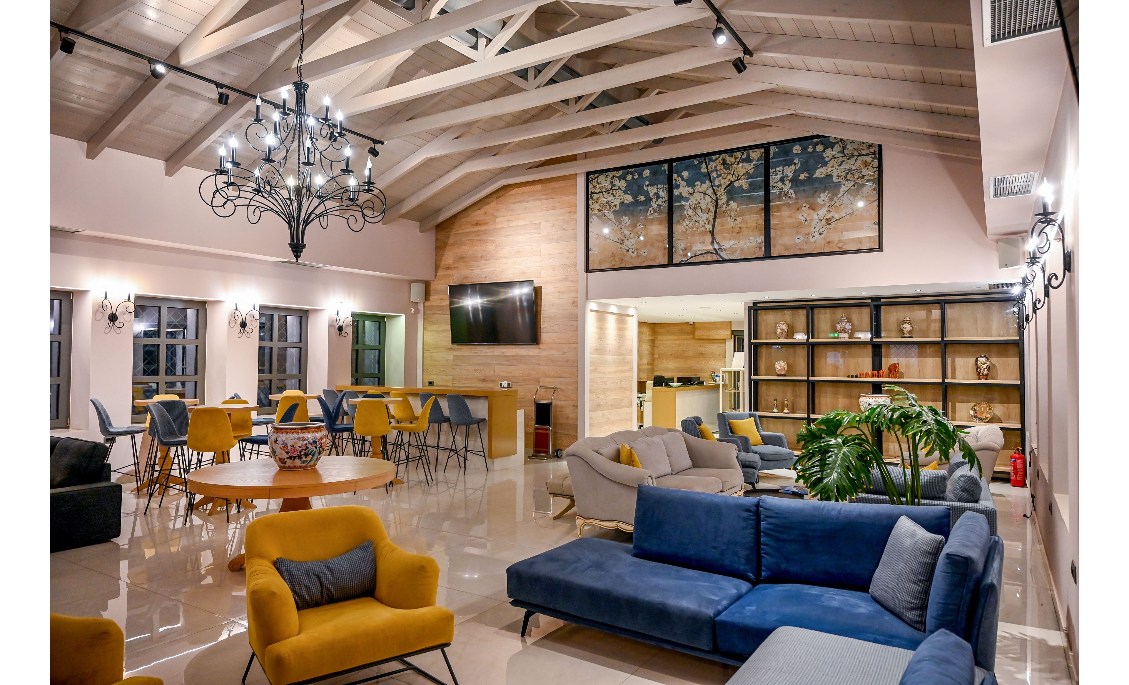 Spacious lounge interior featuring contemporary sofas and armchairs in blue and mustard yellow. The room has high vaulted ceilings with exposed beams and a decorative chandelier, creating an inviting social space. A large wall-mounted television, round wooden table, and open shelving with decor items add functional and aesthetic interest. Warm lighting and polished floors enhance the cozy yet modern atmosphere, complemented by indoor plants and framed wall art.