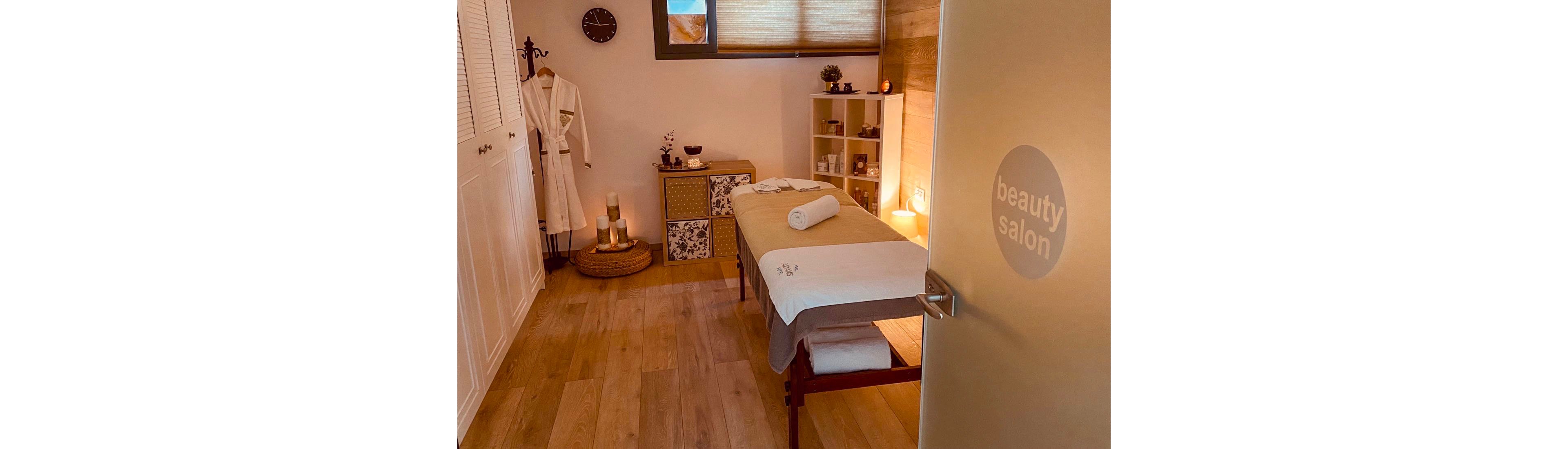 A warm, inviting beauty salon treatment room features a massage table neatly prepared with folded towels. Soft lighting, candles, and decorative storage units create a relaxing spa atmosphere. A robe hangs near a wicker tray with candles, enhancing the sense of comfort and self-care. The wooden floor and neutral tones contribute to the tranquil, wellness-focused setting.