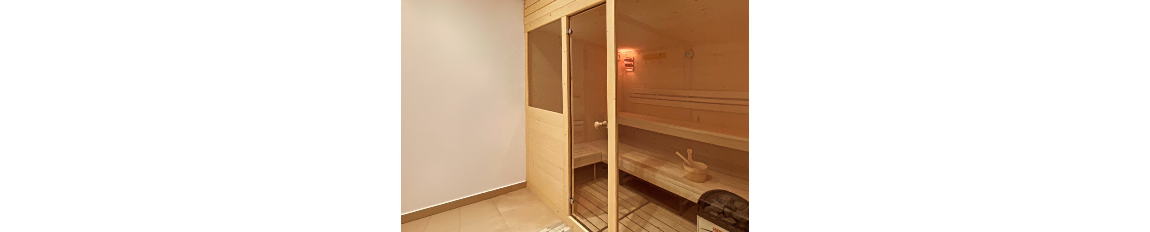 A contemporary wooden sauna cabin stands in a clean indoor space. The sauna features a glass door, wooden benches, and a bucket with a ladle resting on the seating area. Warm ambient lighting glows from inside the cabin, contrasting with the neutral tiled floor and plain wall outside. A heater unit is visible near the entrance, emphasizing the functional spa environment.