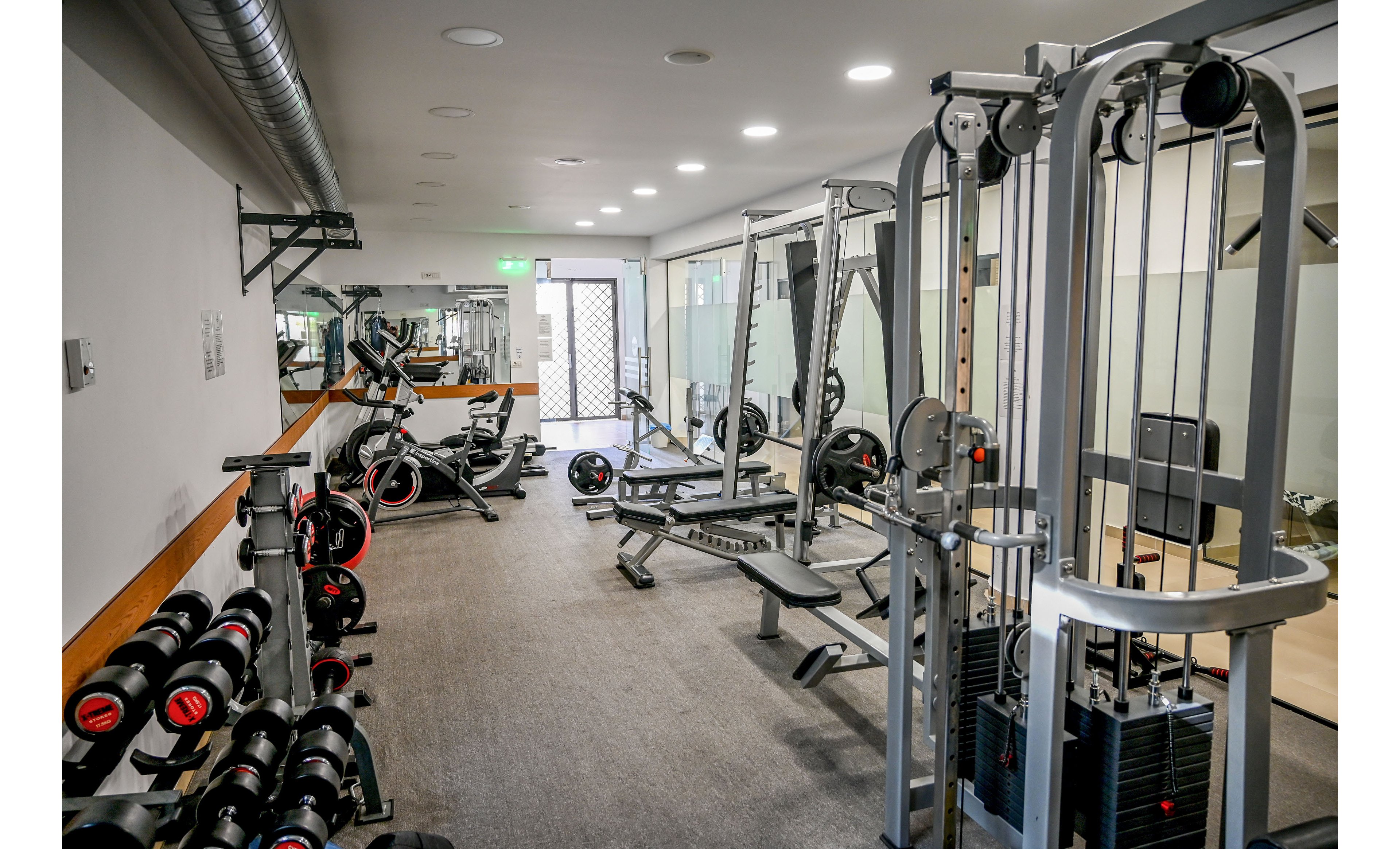 A clean, modern gym interior features a variety of strength training machines and free weights. Dumbbells are neatly arranged on racks along the wall, with benches and resistance machines positioned on a carpeted floor. Cable machines and weight stacks stand near glass partitions, giving the space an open, bright feel. The room is empty, emphasizing the organized layout and professional fitness environment.