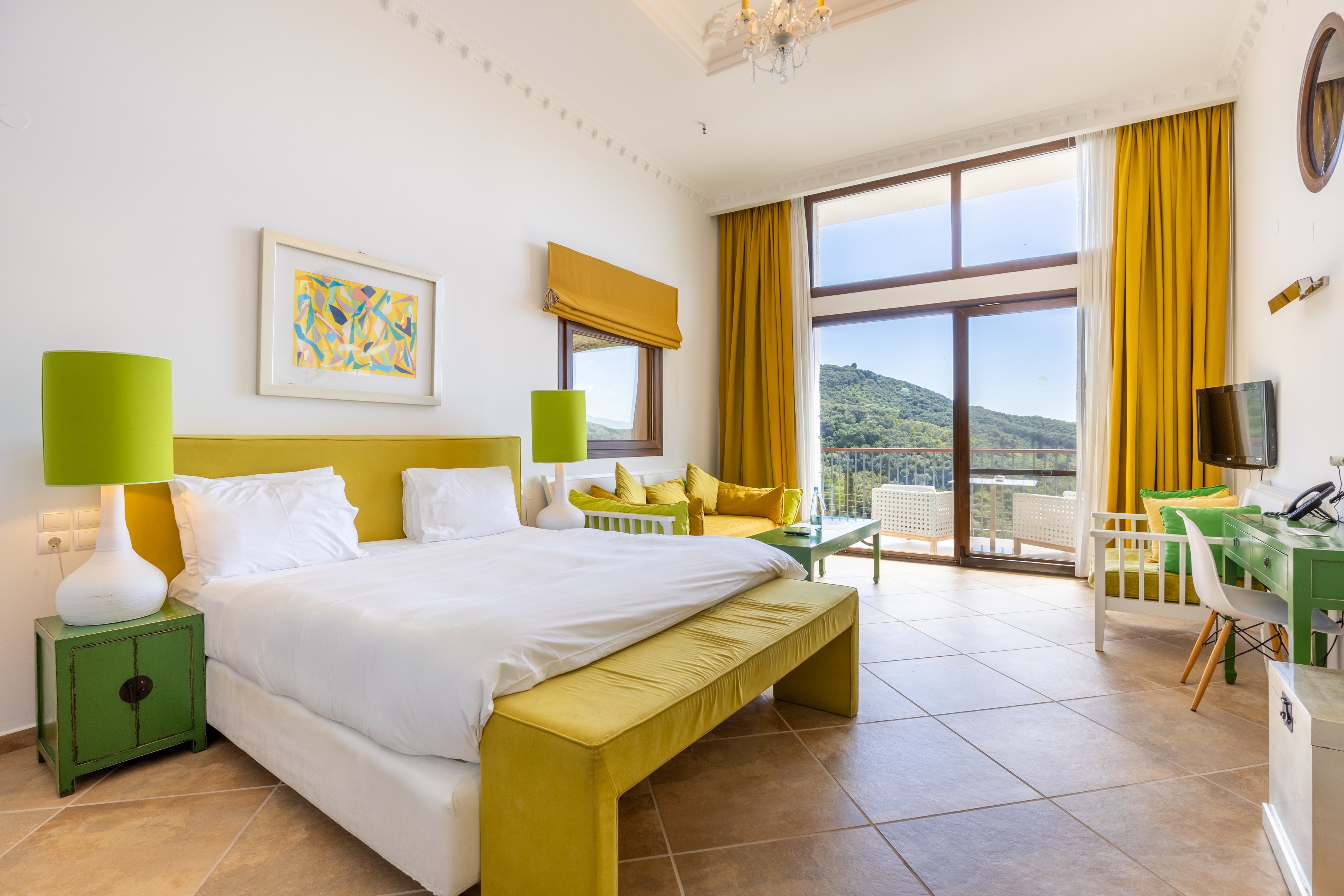 A spacious hotel bedroom features a large double bed with white linens and a yellow upholstered frame. Green bedside lamps and matching side tables add bold color accents to the room. Floor-to-ceiling windows with yellow curtains open to a balcony overlooking green hills and a clear sky. A small seating area, desk with chair, and wall-mounted television complete the modern, inviting interior.