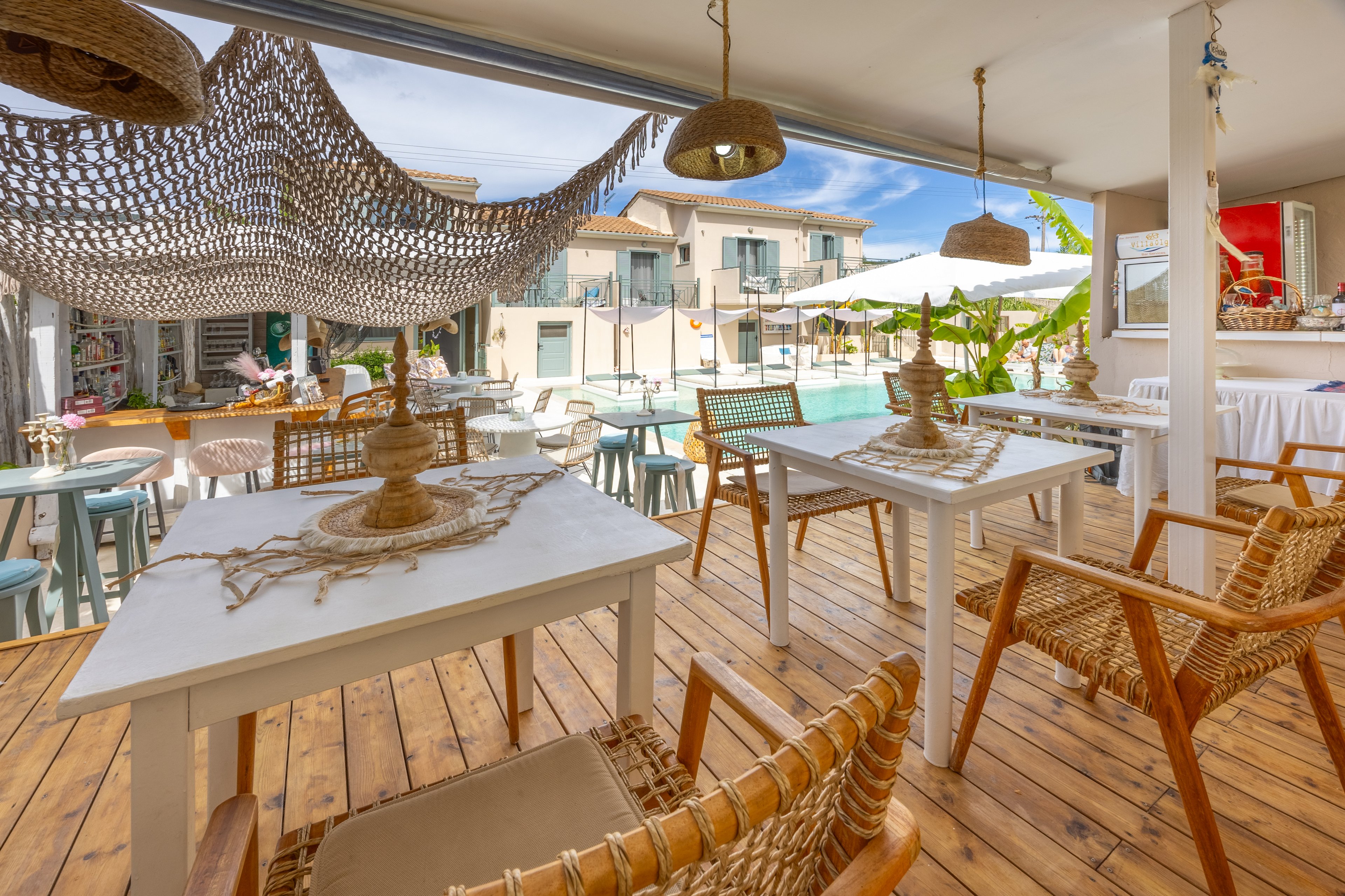 Open-air restaurant terrace with wicker chairs and white tables overlooks a bright swimming pool. Natural wood flooring and woven decor create a relaxed coastal atmosphere. Sunlight filters through a decorative net canopy, casting soft shadows across the seating area. In the background, guests enjoy the pool and surrounding resort buildings under a blue sky.