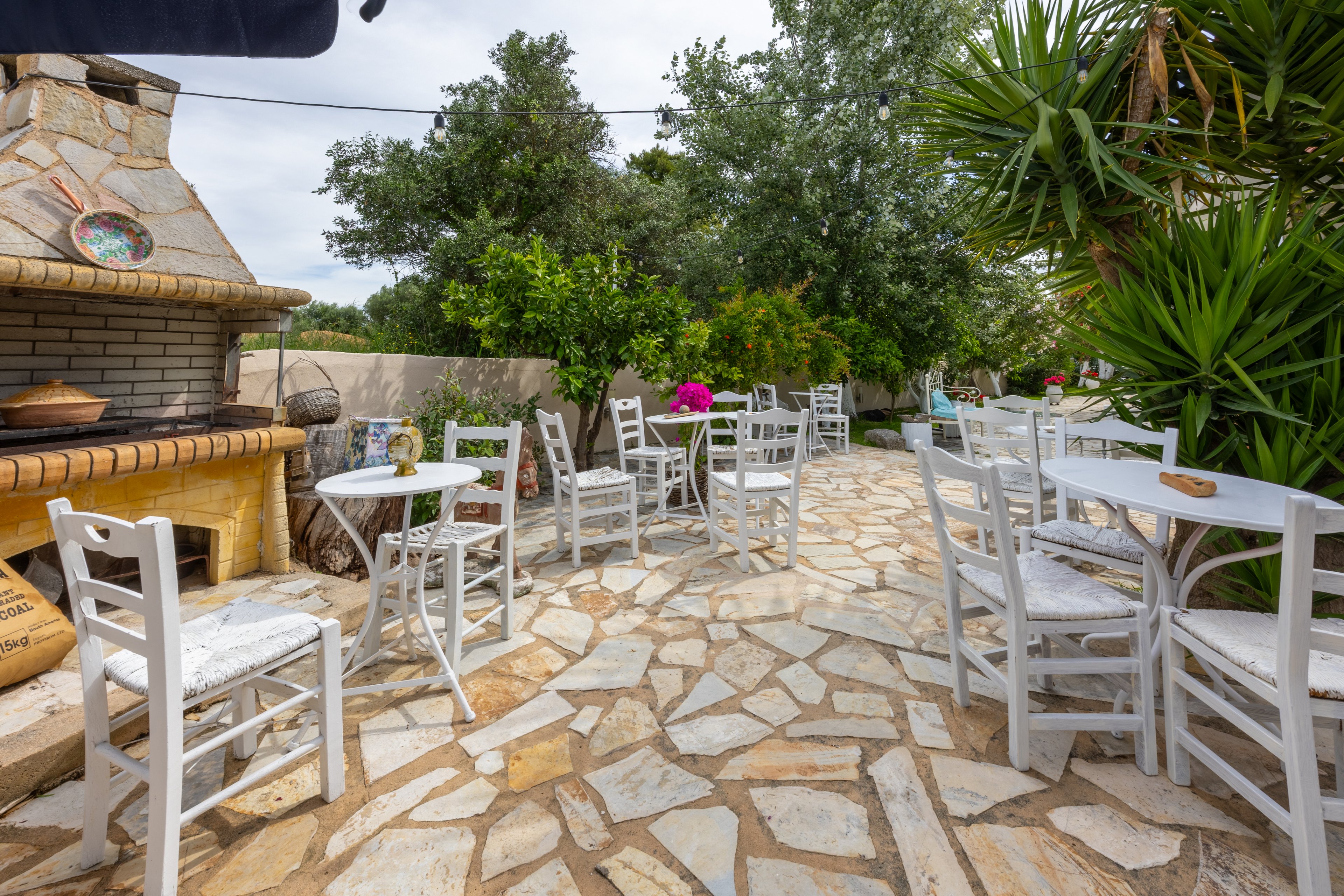An inviting outdoor patio features white wooden chairs and small round tables arranged along a stone-paved terrace. A rustic brick and stone oven with decorative plates and pottery stands to the side, adding a traditional Mediterranean touch. Lush green trees, shrubs, and potted flowers surround the seating area, creating a relaxed garden atmosphere. The scene suggests a quiet café or backyard dining space ideal for leisurely meals and conversation.