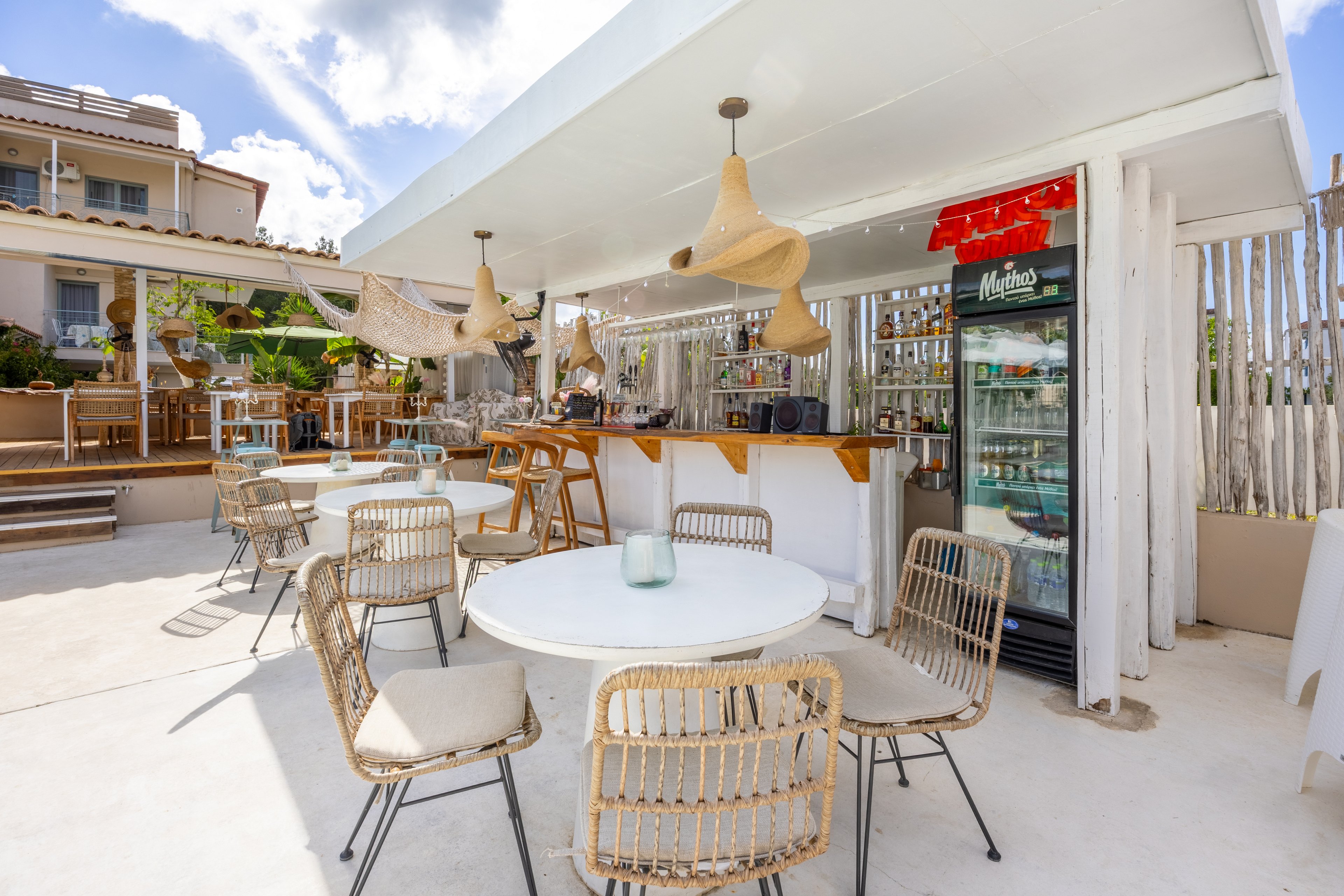 Bright outdoor bar area with round white tables and wicker chairs arranged on a sunlit terrace. A white bar counter with wooden accents stands under a modern canopy, featuring hanging woven pendant lights. A glass-front refrigerator filled with drinks is visible beside the bar, with a red awning above it. The relaxed coastal setting suggests a casual place for guests to enjoy refreshments in warm weather.
