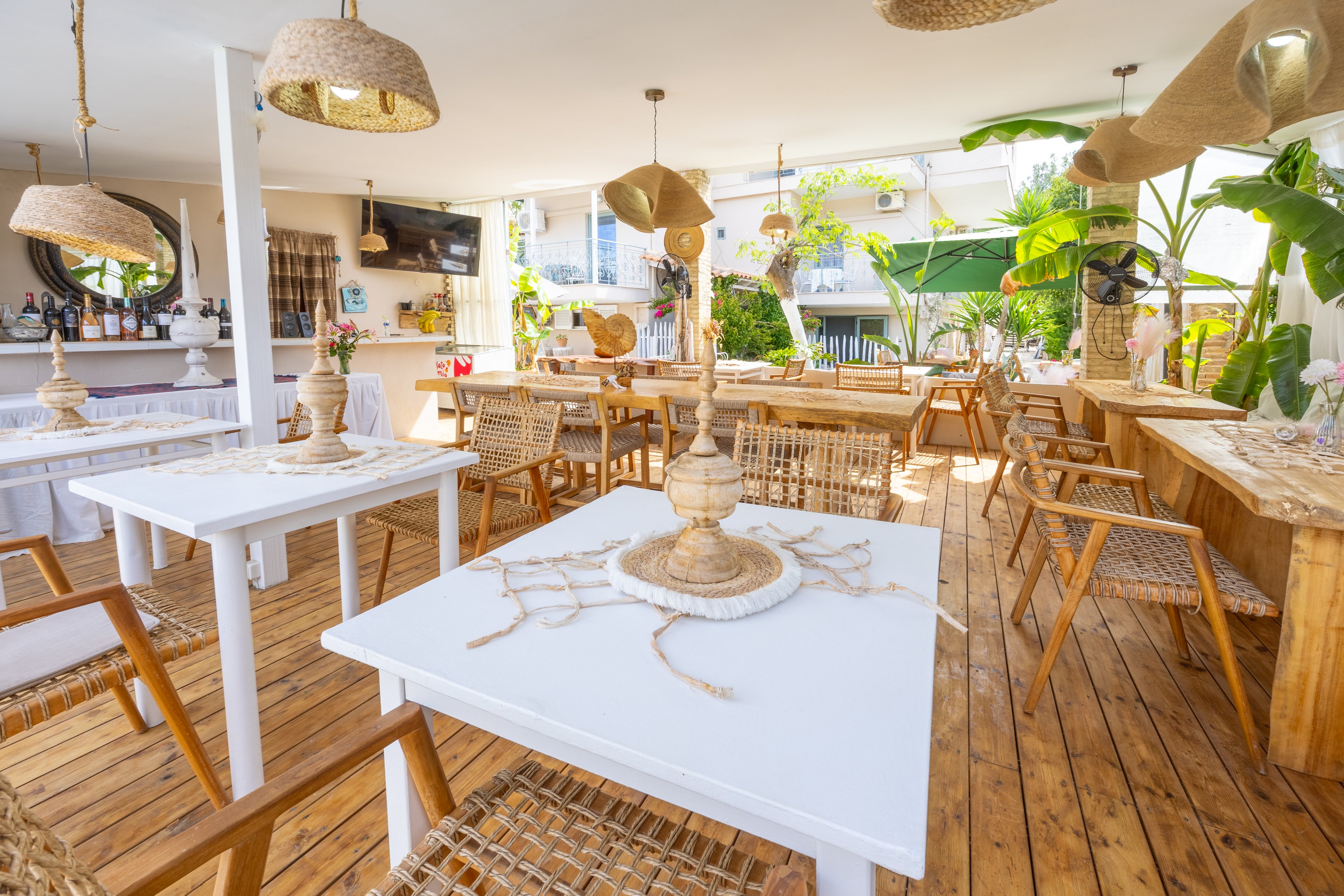 Sunlit open-air restaurant interior featuring white tables and woven wicker chairs on a wooden floor. Natural fiber pendant lamps hang from the ceiling, casting warm light across the space. Decorative rope centerpieces and plants add a relaxed coastal touch. A bar area with bottles and glassware is visible in the background, enhancing the casual dining atmosphere.