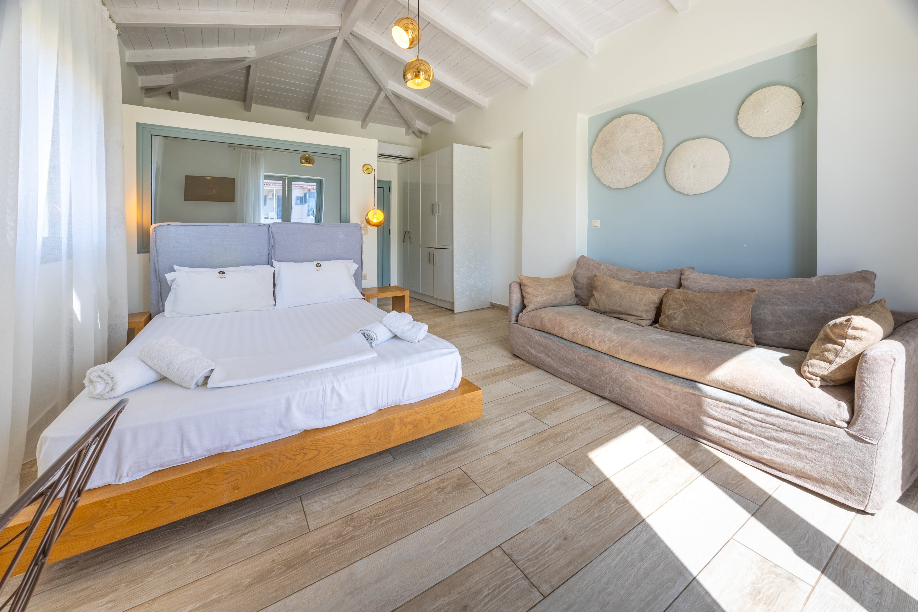 A sunlit bedroom features a low wooden platform bed neatly made with white linens and towels. To the right, a large beige sofa provides additional seating in the airy space. Light wood flooring and white beamed ceilings enhance the bright, coastal feel of the room. Soft blue accent walls with round wall art and warm pendant lights add subtle decorative touches.