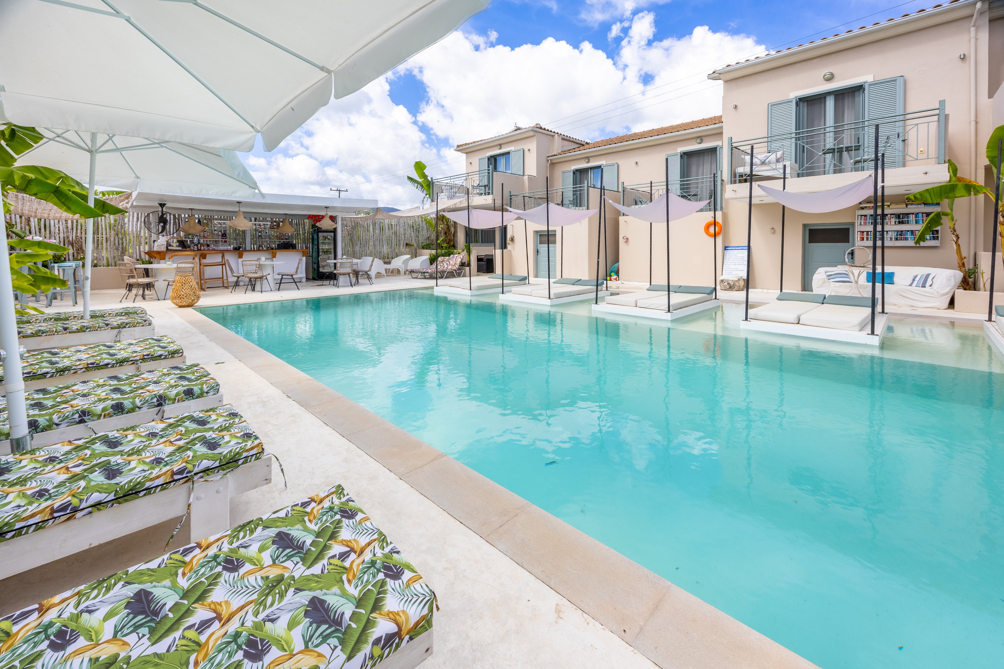 A serene outdoor swimming pool is surrounded by patterned sun loungers and shaded cabana-style daybeds. The scene appears to be part of a modern boutique hotel or resort with light-colored buildings and balconies overlooking the water. White umbrellas and tropical-print cushions add a relaxed, vacation atmosphere. The bright daylight and blue sky with scattered clouds enhance the fresh, inviting feel of the pool area.