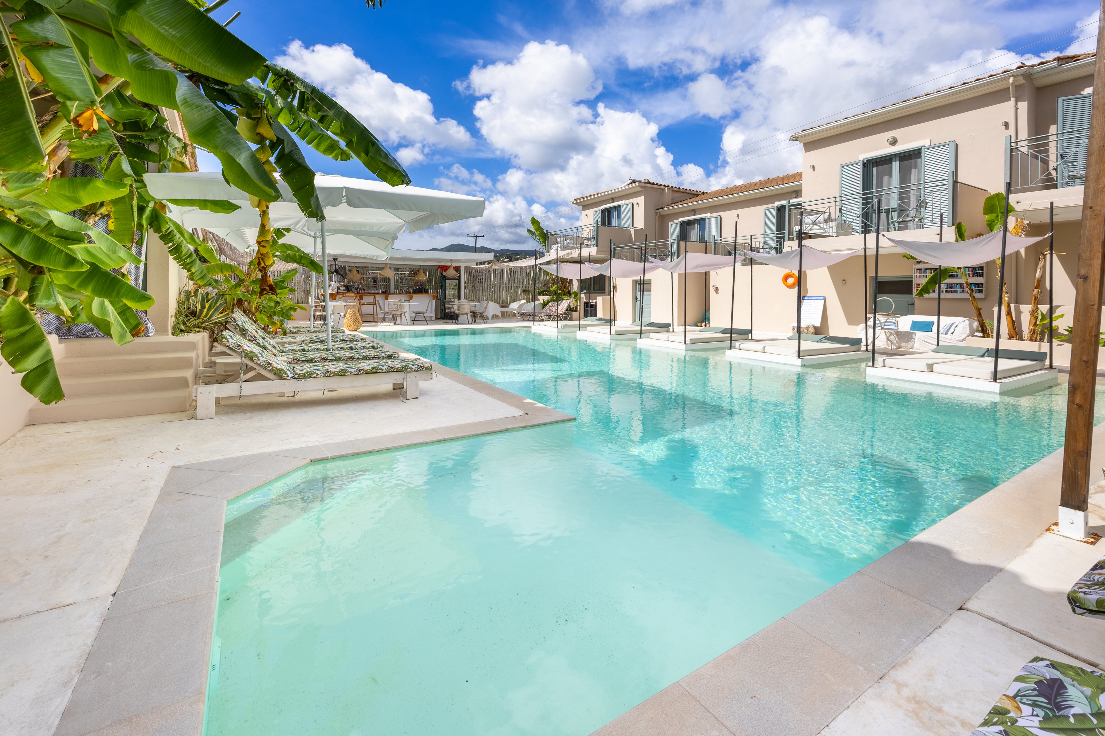 A long turquoise swimming pool stretches between modern beige resort buildings under a bright blue sky with scattered clouds. Sun loungers and umbrellas line the poolside, creating a relaxing vacation atmosphere. Banana plants and light-colored stone surfaces add a tropical, contemporary touch. The scene is calm and inviting, suggesting a sunny day at a stylish hotel or holiday apartment complex.