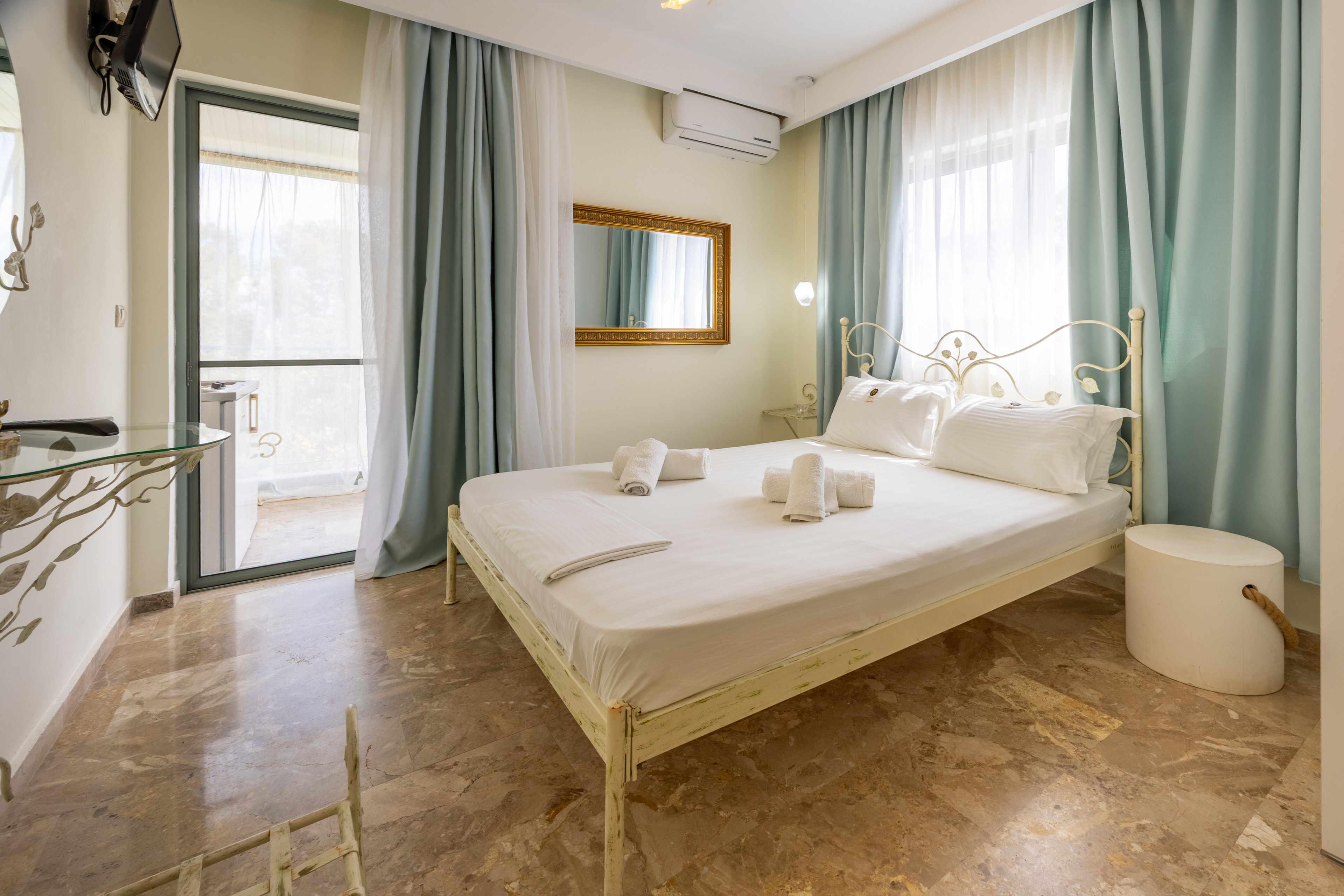 A sunlit bedroom features a white metal bed neatly made with white linens and rolled towels. Light blue and white curtains frame a large window and glass door leading to a balcony. A wall-mounted mirror, small side table, and mounted television add functional details to the space. The room has a polished stone floor and a soft, neutral color palette that feels calm and inviting.