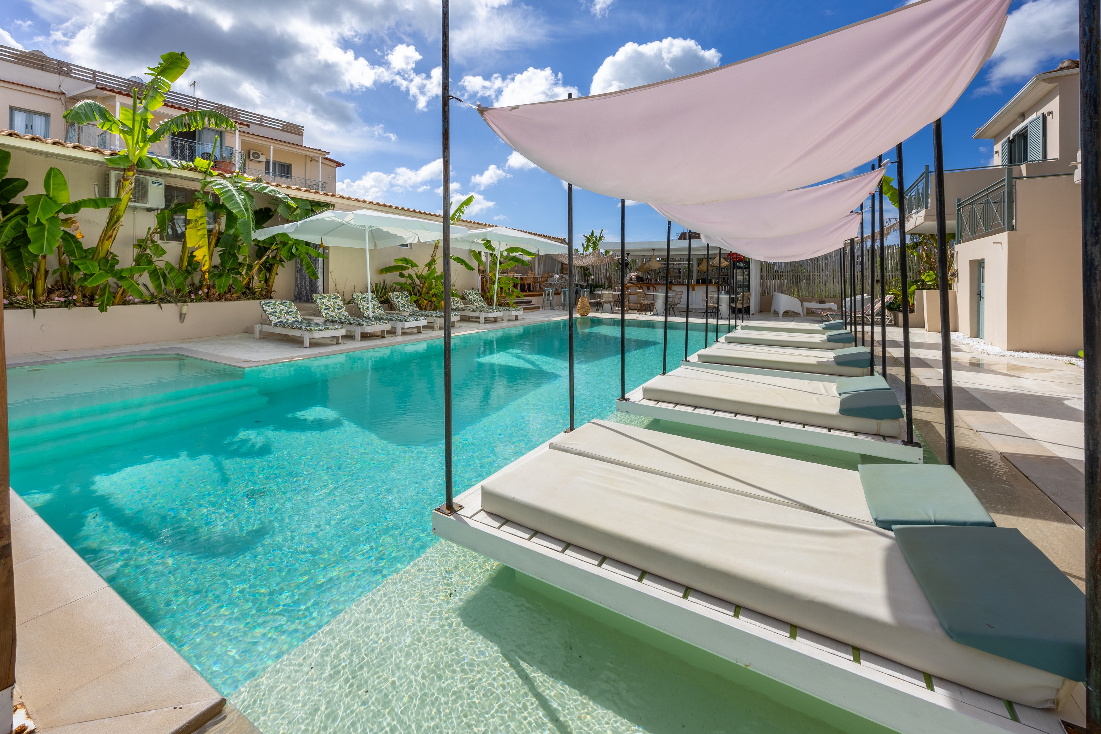 A contemporary resort swimming pool features a row of low loungers partially submerged in shallow water. White fabric canopies hang above the loungers, creating shaded relaxation areas along the poolside. Tropical plants and palm trees line the far side, with additional sunbeds and umbrellas visible in the background. Surrounding buildings and a bright blue sky with scattered clouds complete the tranquil vacation setting.