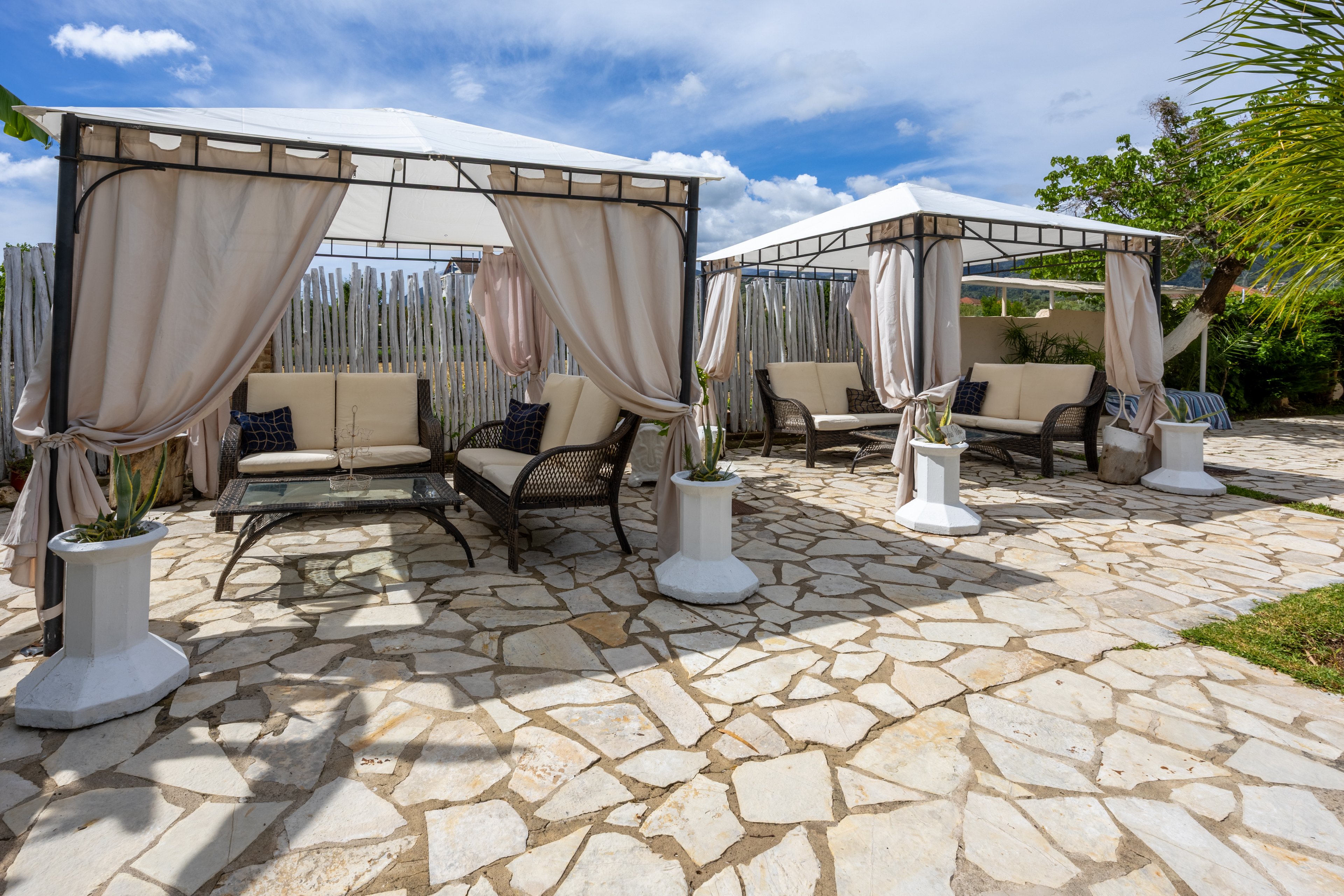 An outdoor lounge area features several cabanas with flowing beige curtains and cushioned wicker seating. The cabanas are arranged on a stone-paved patio under a bright blue sky with scattered clouds. Potted plants sit on white pedestal stands beside the seating areas, adding greenery to the scene. A wooden fence and surrounding trees frame the relaxing, resort-like setting.
