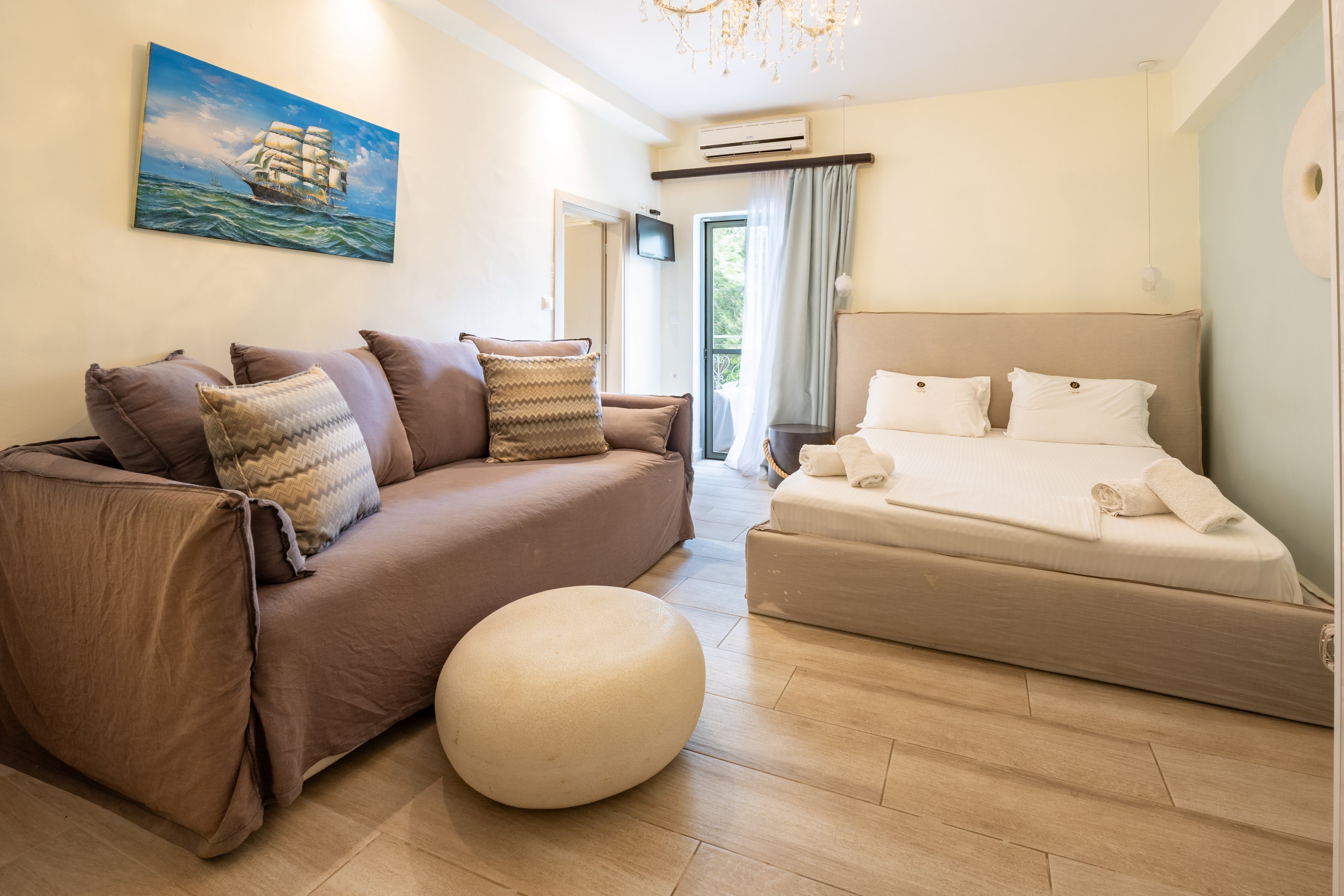 A bright, modern hotel room features a large beige bed neatly made with white linens and towels. A comfortable sofa with patterned cushions sits opposite, accompanied by a round ottoman on a light wooden floor. Natural light enters through a glass door leading to a balcony with greenery outside. Soft neutral tones, a chandelier, and a nautical ship painting on the wall create a calm, inviting atmosphere.