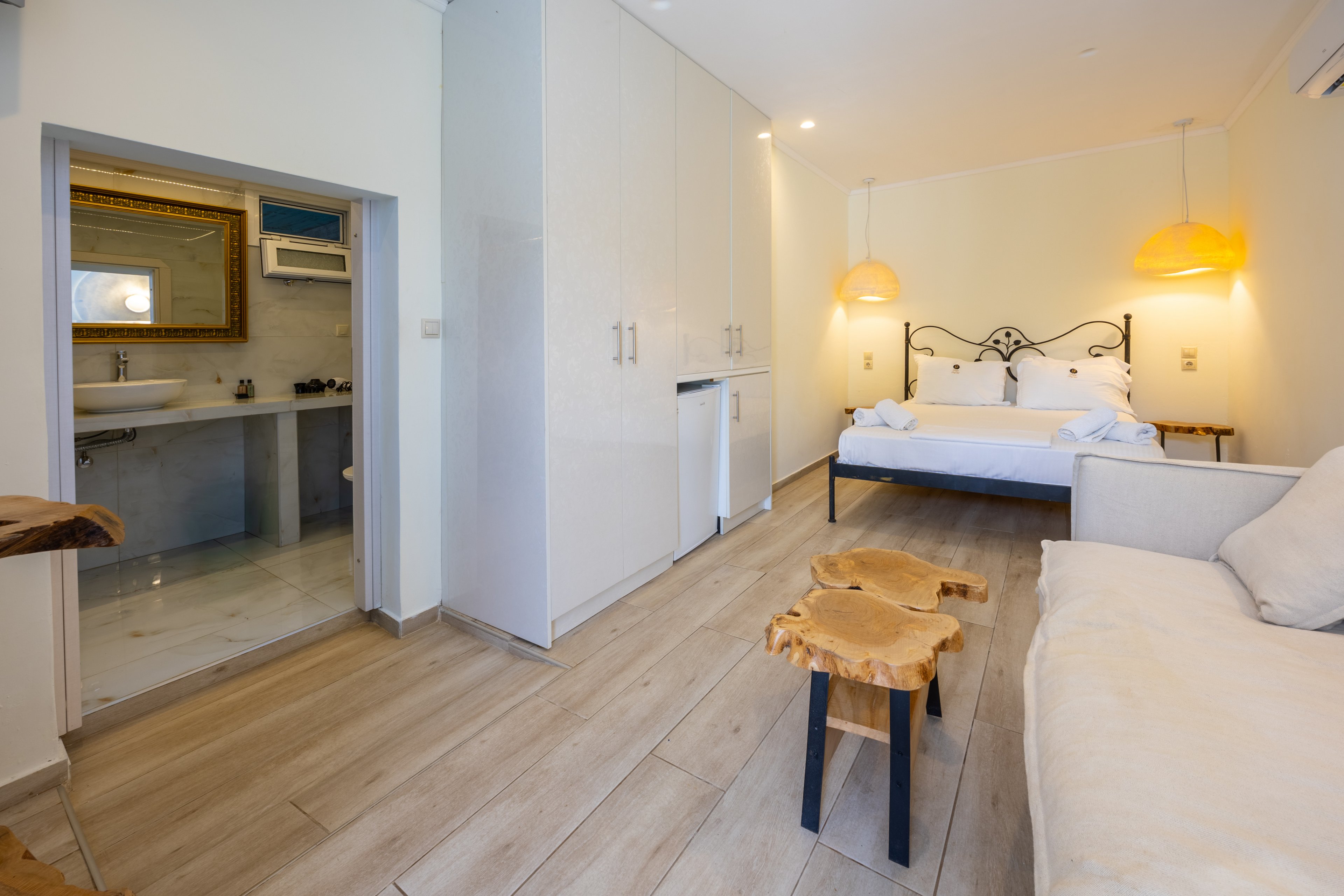 A bright, modern hotel room features a double bed with white linens and warm hanging lamps. Light wood flooring and white cabinetry create a clean, minimalist aesthetic. A small sofa and unique wooden side tables add comfort and character to the space. An open doorway reveals an ensuite bathroom with a large mirror and countertop.