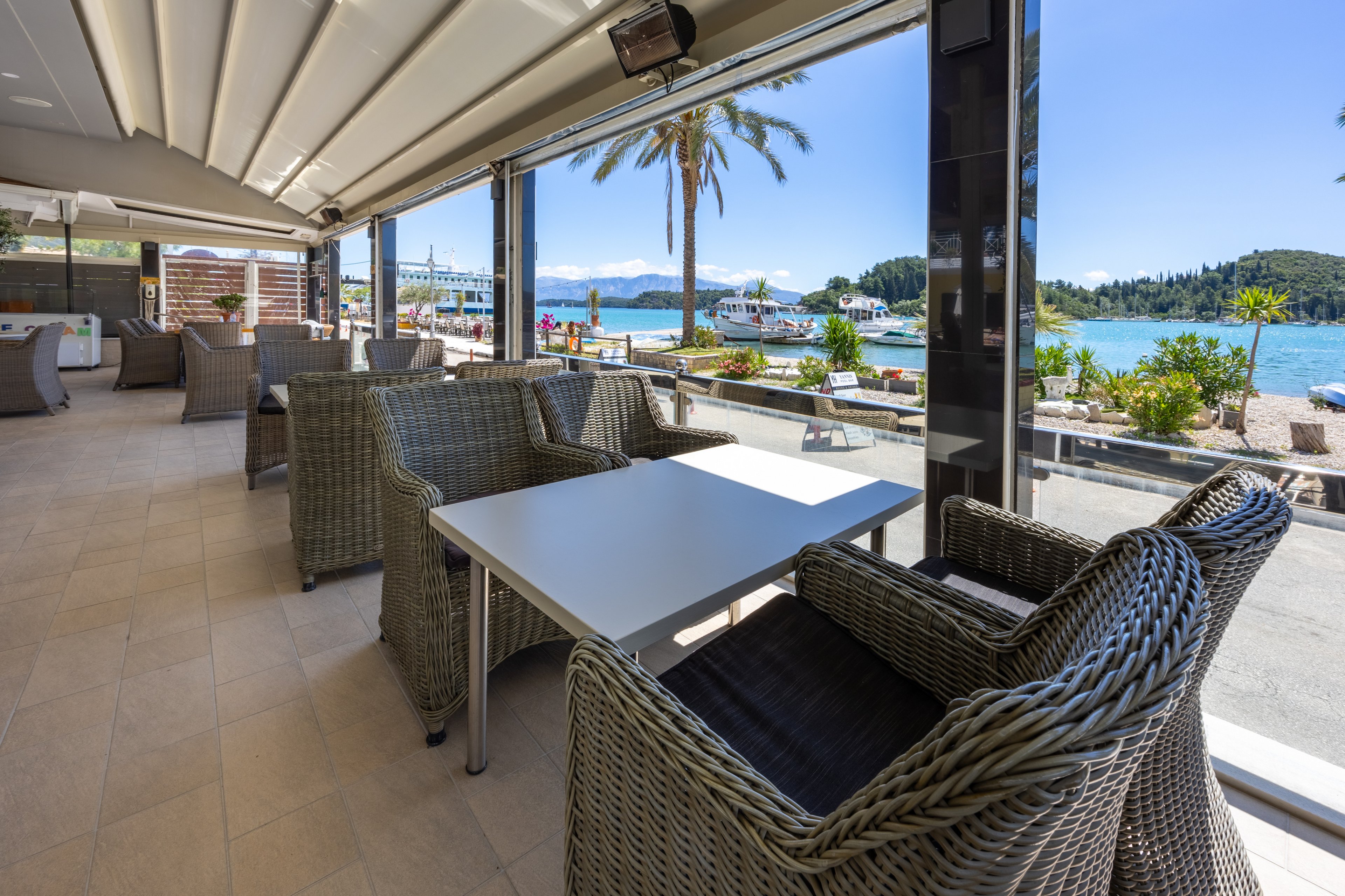 An open-air cafe terrace features wicker armchairs around a white table overlooking a scenic waterfront. Large glass walls and a retractable roof create a bright, airy dining space. Boats are docked along the pier, with blue water, distant islands, and mountains in the background. A tall palm tree and potted plants add a relaxed, coastal atmosphere under clear sunny skies.