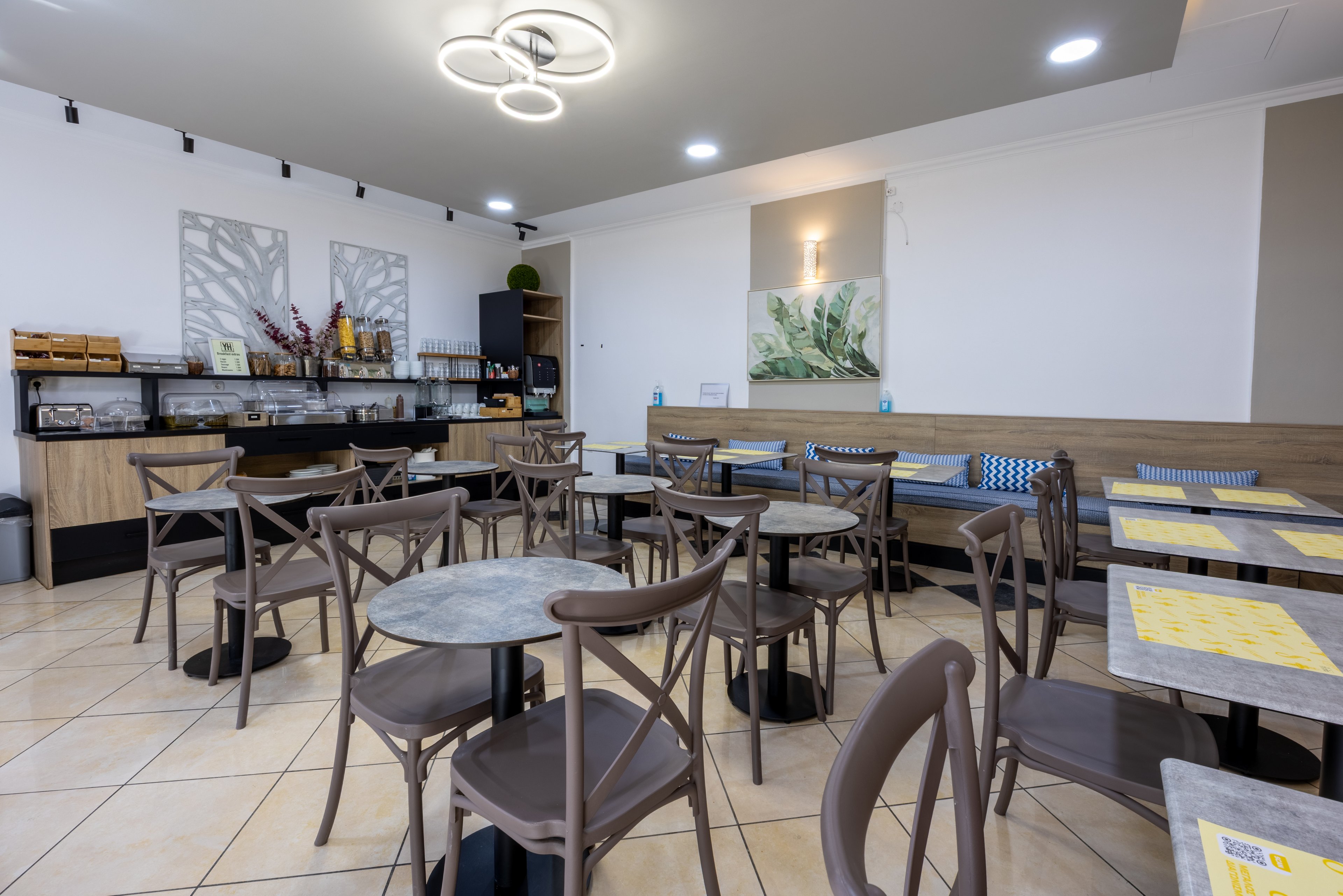 A clean, modern cafe dining area features multiple small tables with matching chairs arranged neatly across a tiled floor. The space is empty of people, emphasizing the orderly layout and ample seating. A buffet-style counter with coffee machines, cups, and food containers lines the back wall. Contemporary ceiling lights and nature-themed wall art contribute to a bright, welcoming atmosphere.