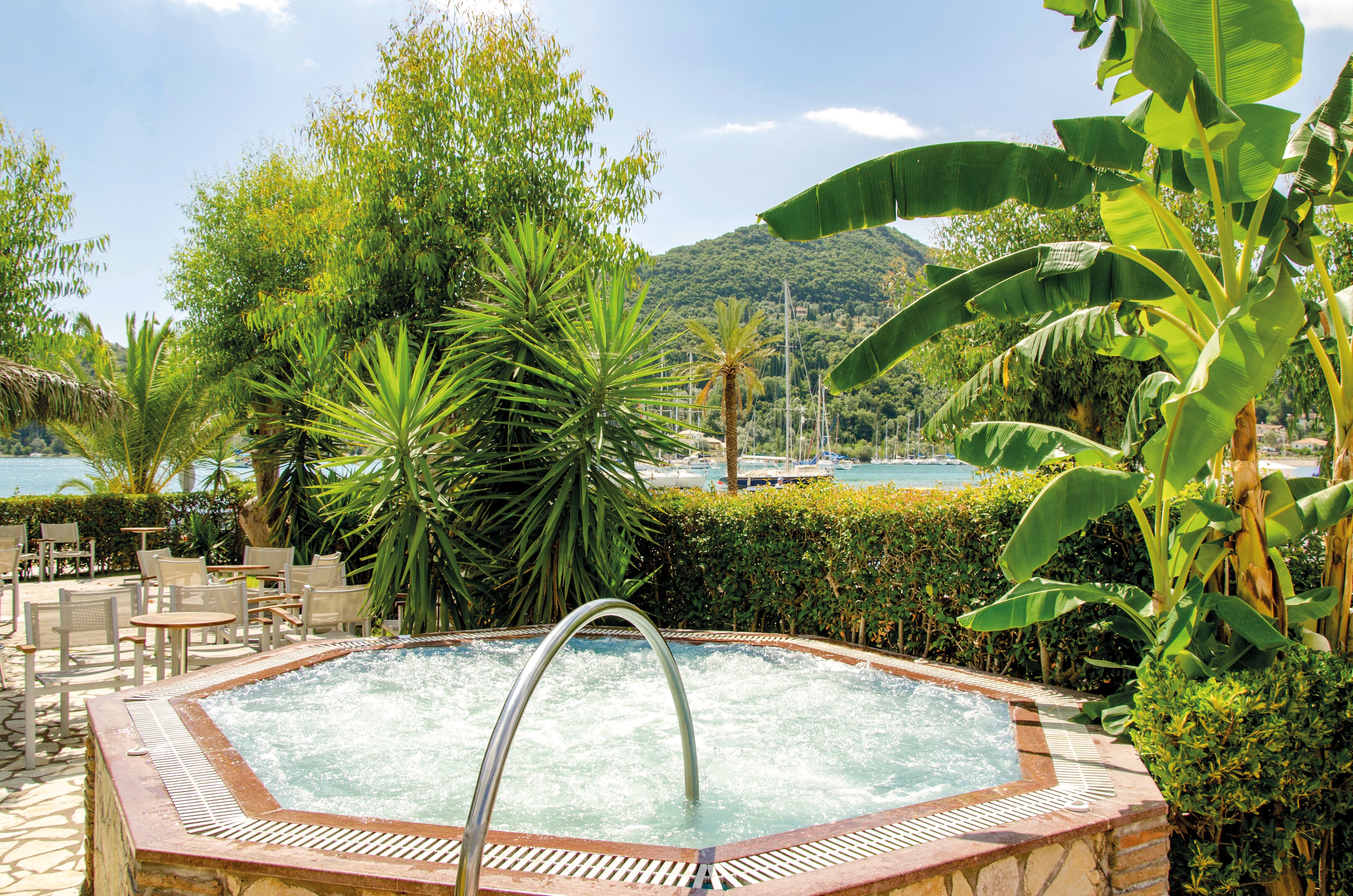 An inviting outdoor hot tub surrounded by lush tropical plants, including banana trees and palms. The setting features a serene backdrop of a marina with sailboats and green hills. The scene is bright and sunny, evoking relaxation and leisure in a resort-like atmosphere.