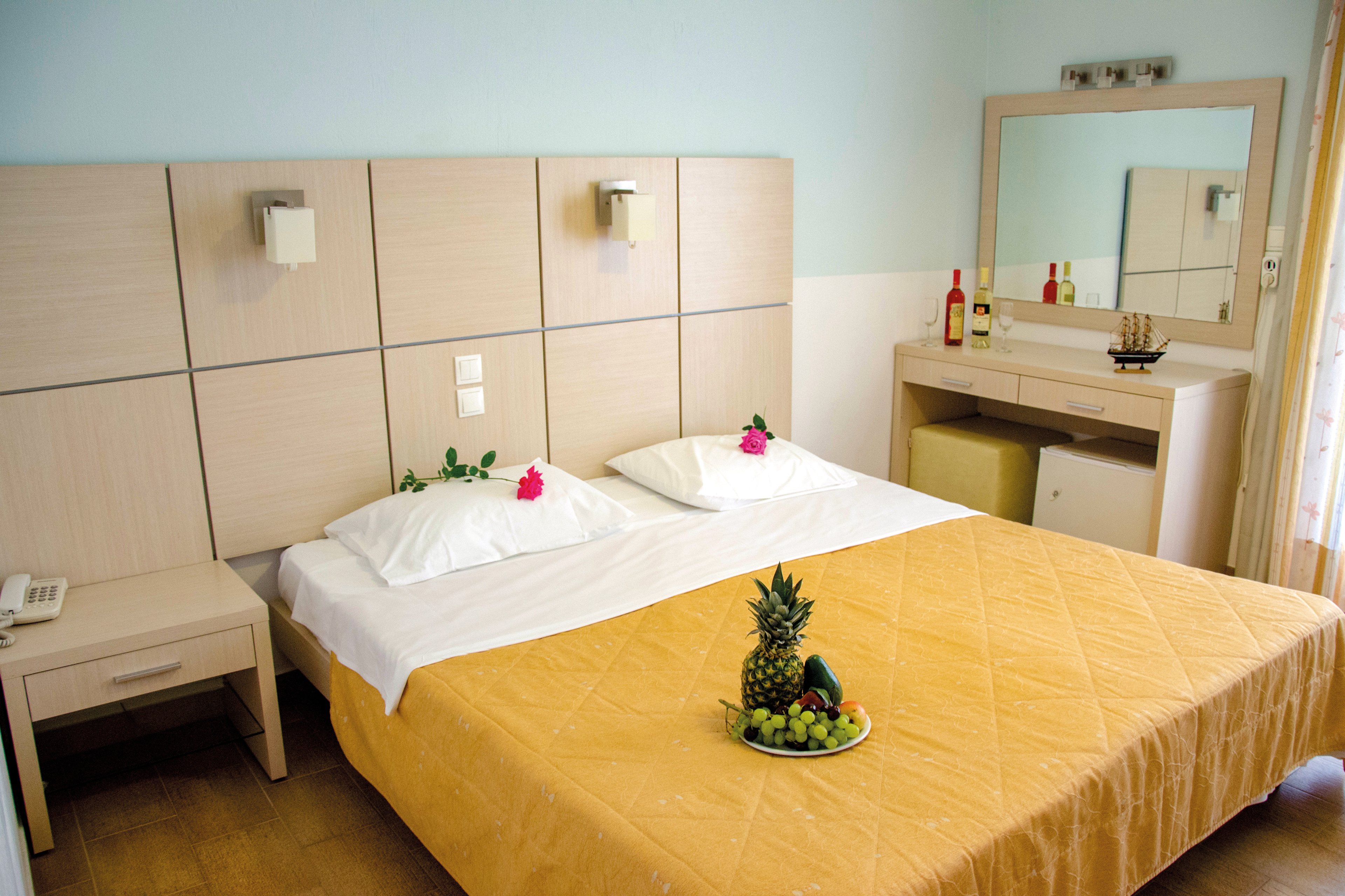 A well-lit hotel room featuring a double bed with white linens and a yellow quilt. The room includes a wooden nightstand, a mirror, and a dresser with two bottles of wine and decorative items. A fresh fruit platter with pineapple, grapes, and other fruits is placed on the bed. The setting is clean and inviting, ideal for relaxation.