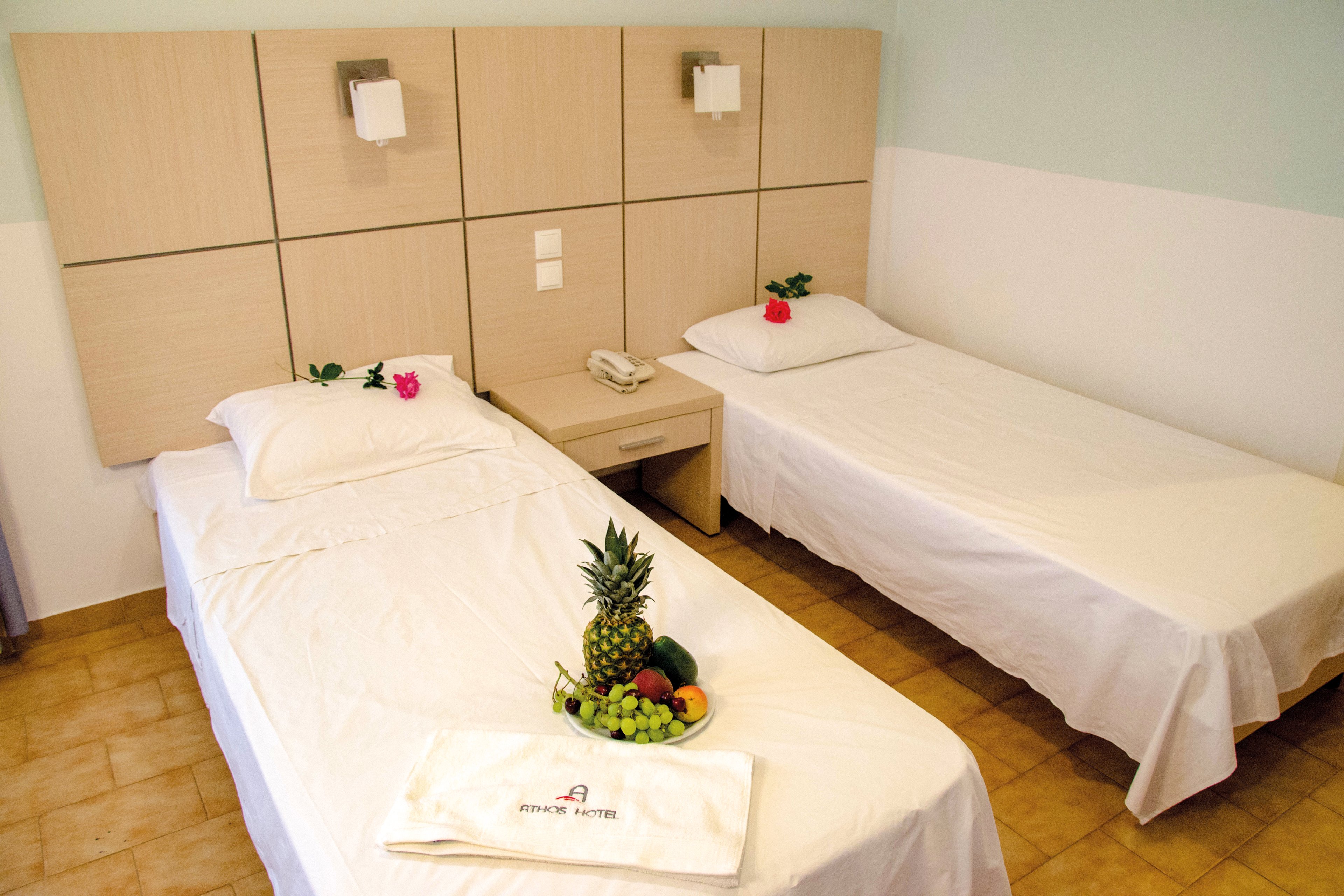 A neatly arranged hotel room featuring two twin beds with white linens and decorative pink flowers. A wooden nightstand sits between the beds, holding a telephone and wall-mounted lights above. A fresh fruit platter with pineapple, grapes, and other fruits is placed on a towel labeled 'Prince Hotel'. The room has a clean and minimalistic design with light-colored walls and tiled flooring.