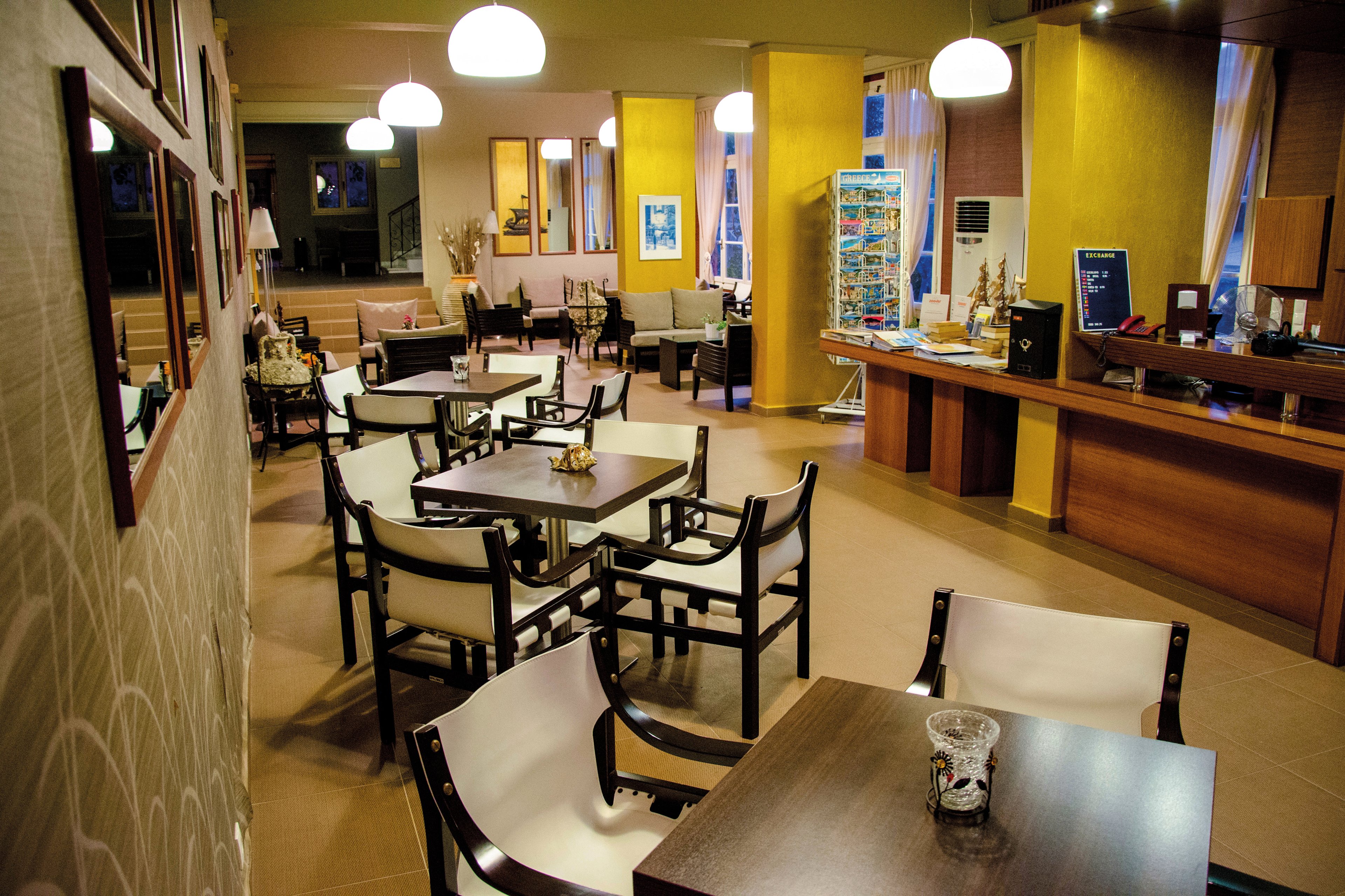 A modern cafe setting featuring wooden tables and chairs arranged neatly in a spacious room. Warm yellow walls and pendant lights create a welcoming ambiance. Visible details include a counter area with a display rack and framed artwork on the walls. The space is designed for casual dining or social gatherings.