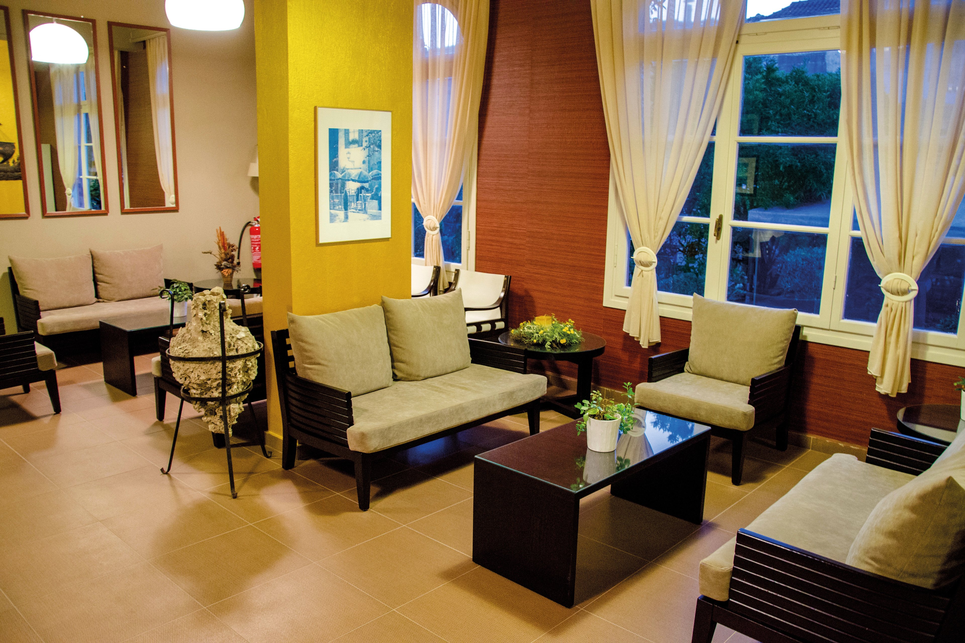 A warm and inviting lounge featuring modern wooden furniture with beige cushions. The setting includes large windows with cream curtains, vibrant yellow and red walls, and decorative plants on tables. The space is well-lit with soft lighting, creating a relaxing atmosphere.