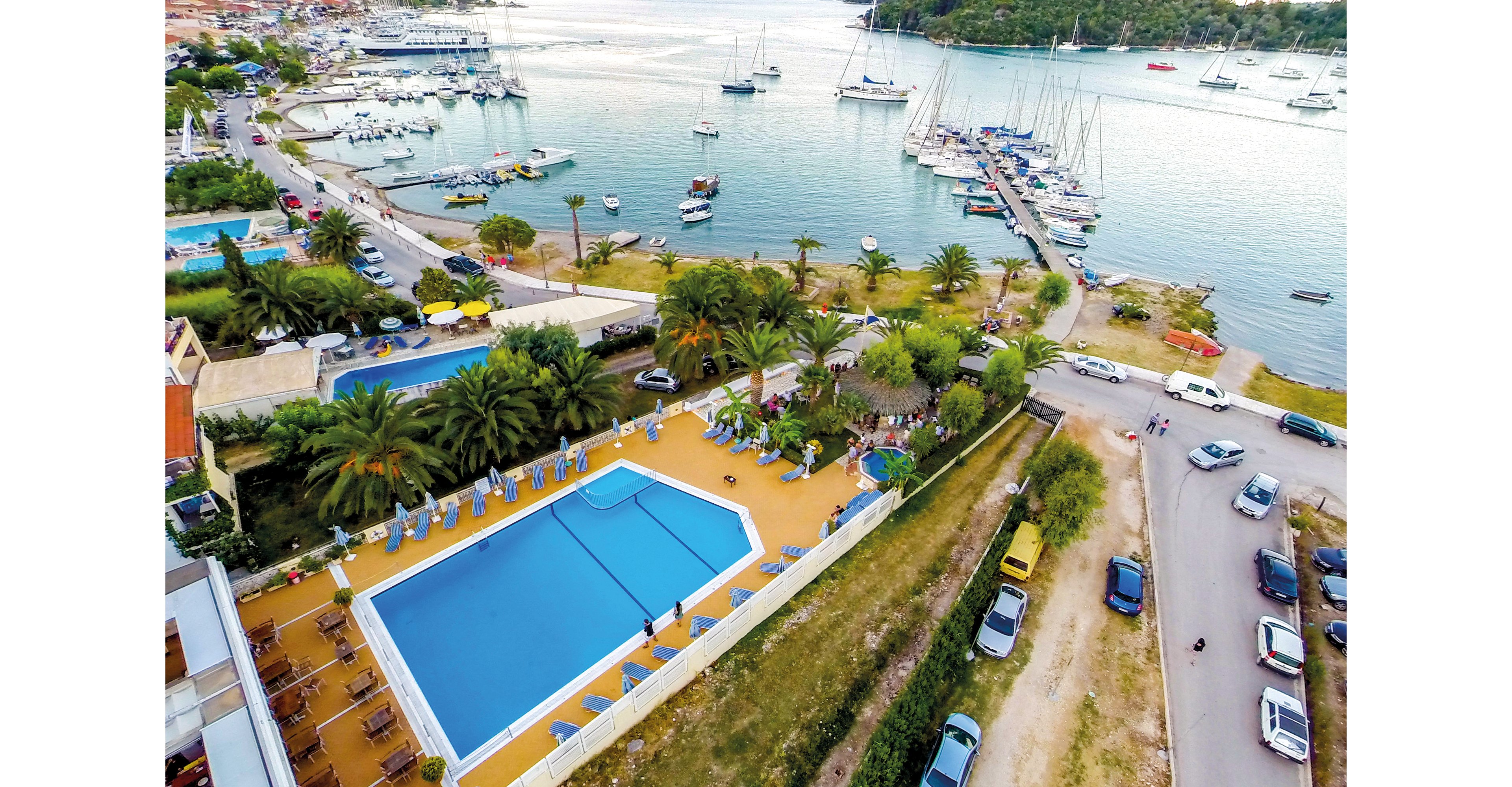 An aerial shot showcasing a large swimming pool surrounded by lounge chairs and palm trees, situated near a marina with docked sailboats. The setting includes a coastal town with roads, parked cars, and lush greenery. The vibrant blue pool contrasts with the earthy tones of the surrounding area, creating a serene vacation atmosphere.