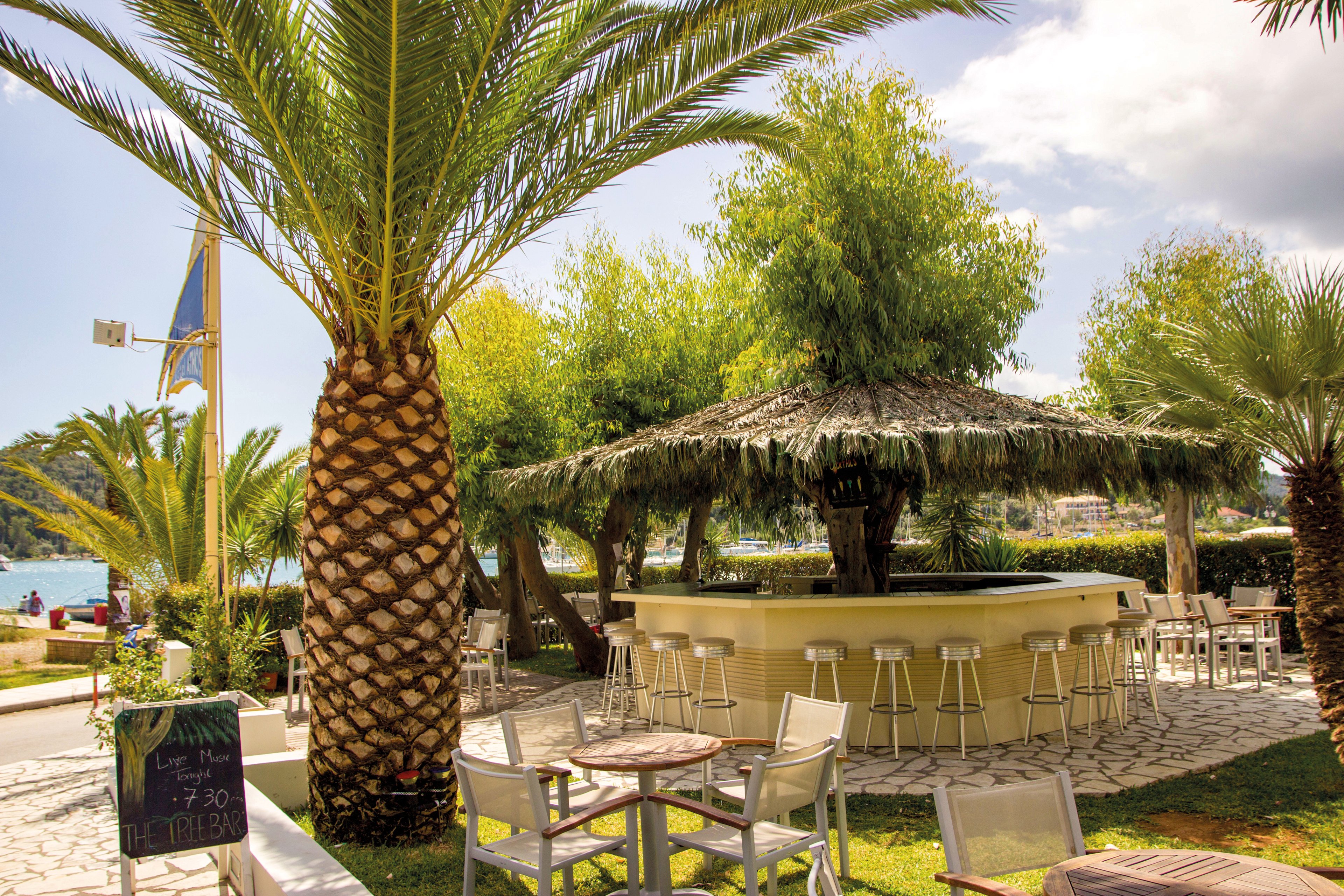 A circular outdoor bar surrounded by lush palm trees and greenery, set in a sunny tropical location. White chairs and tables are arranged around the bar, creating a relaxed atmosphere. A chalkboard sign is visible, displaying the time '17:30'. The setting overlooks a serene waterfront under a partly cloudy sky.