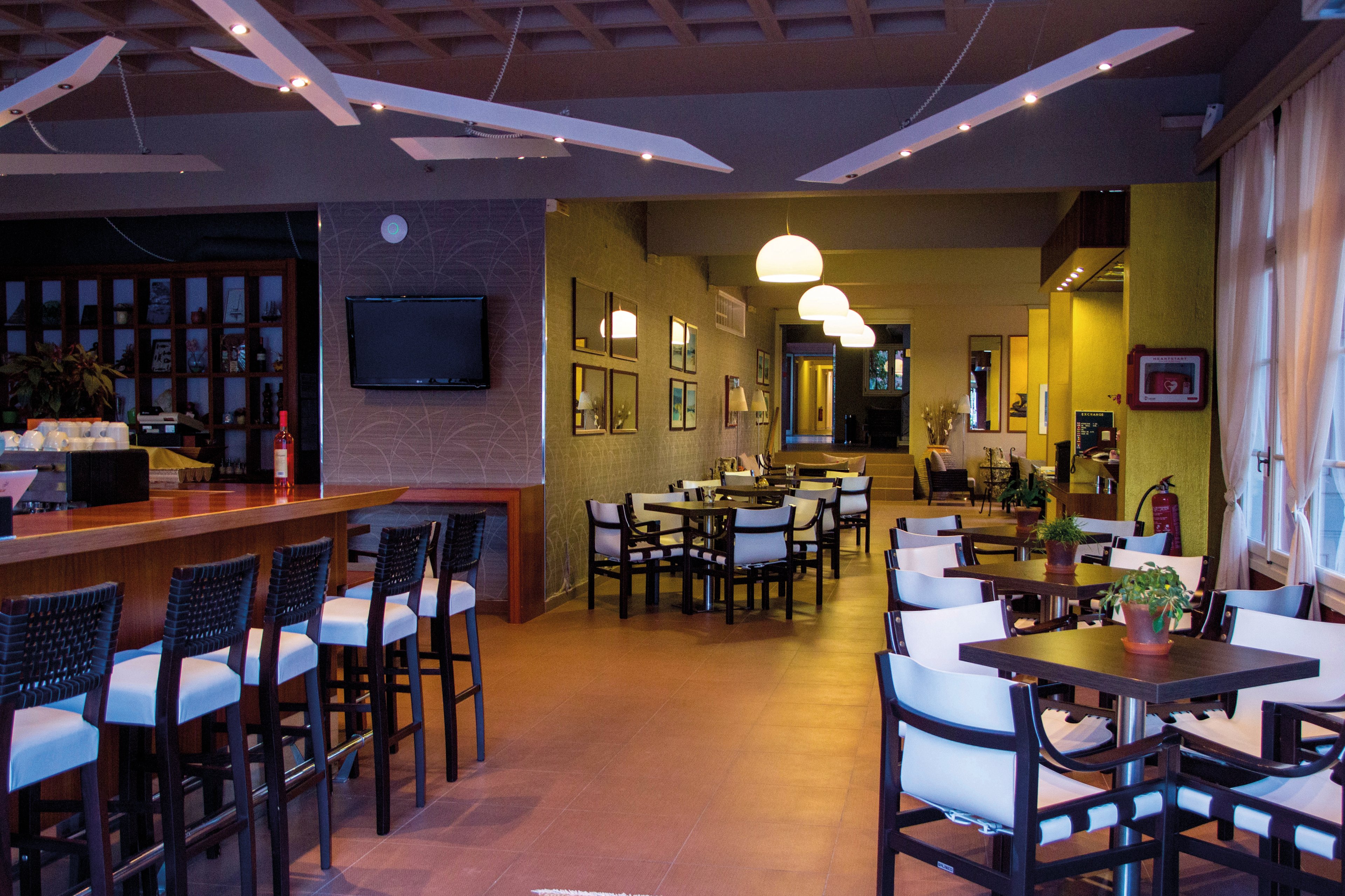 A contemporary restaurant featuring a stylish bar area with high chairs and tables arranged for dining. The setting includes warm lighting, decorative ceiling elements, and indoor plants adding a cozy ambiance. Visible details include a television screen and wall-mounted mirrors enhancing the space.
