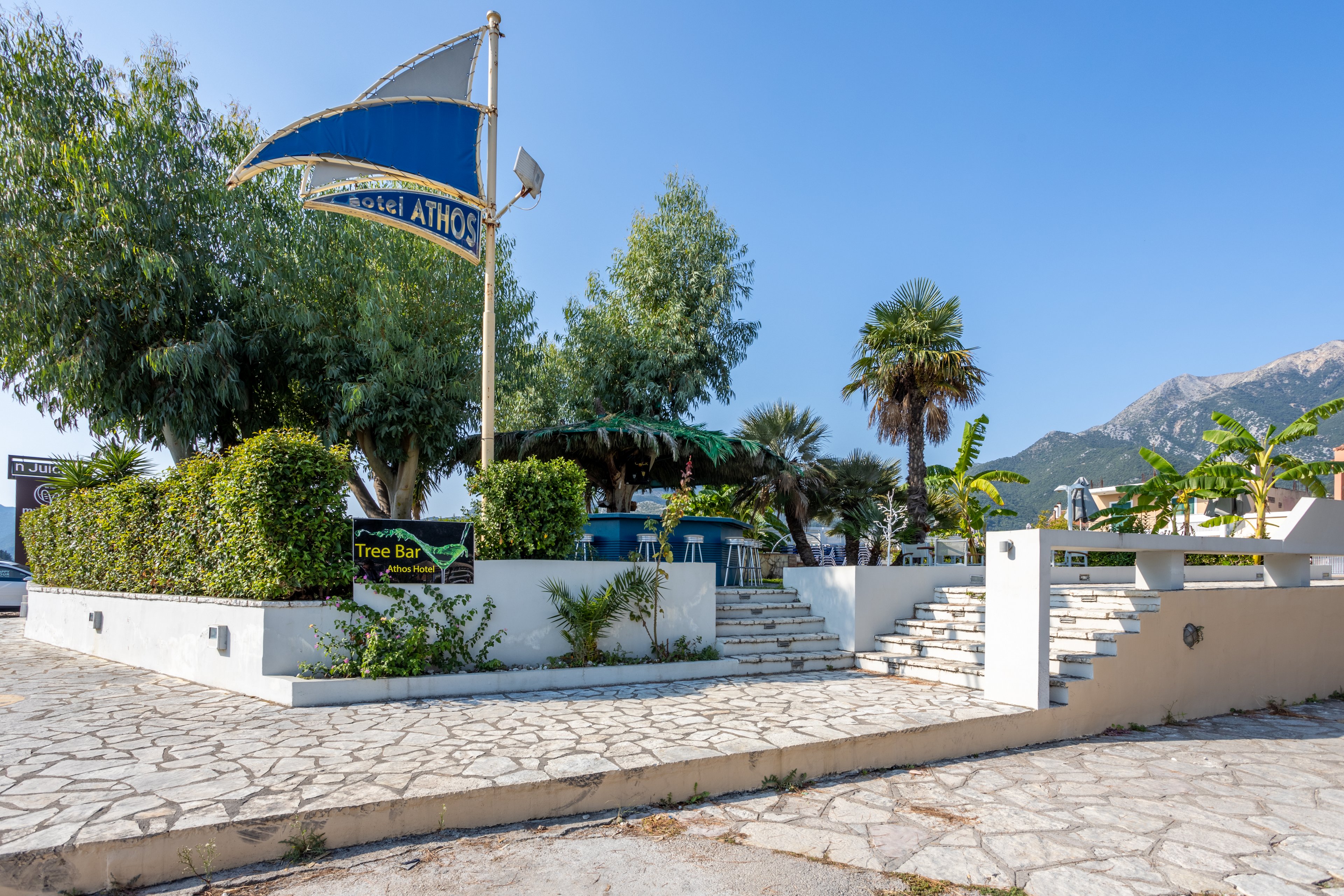 The image showcases the exterior of Hotel Athos, featuring a prominent blue and white flag with the hotel name clearly visible. The setting includes lush tropical plants, a stone-paved walkway, and a staircase leading to a shaded seating area. A sign for 'Tree Bar' is also visible, adding to the inviting atmosphere. The backdrop includes a mountainous landscape under a clear blue sky.