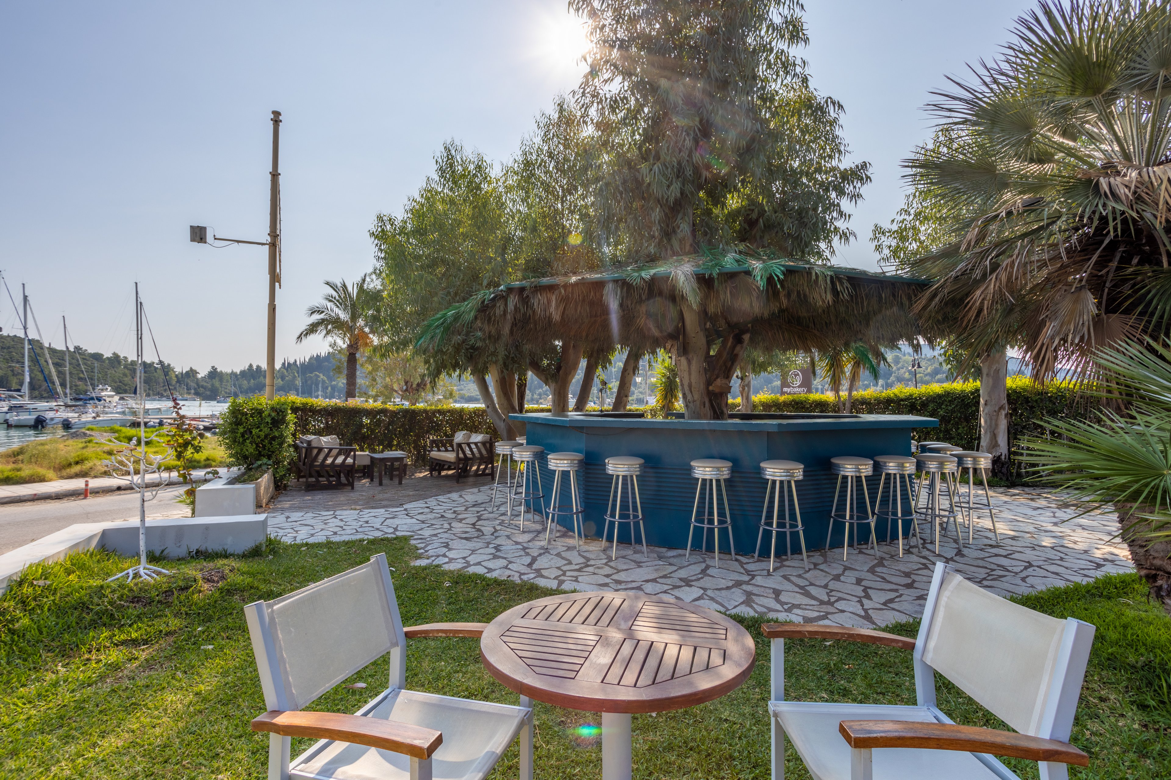 An open-air bar surrounded by lush greenery and palm trees, featuring a circular counter with bar stools. The setting overlooks a marina with docked sailboats under a bright, sunny sky. Wooden chairs and a table are placed on the grass, creating a relaxed atmosphere for outdoor leisure.