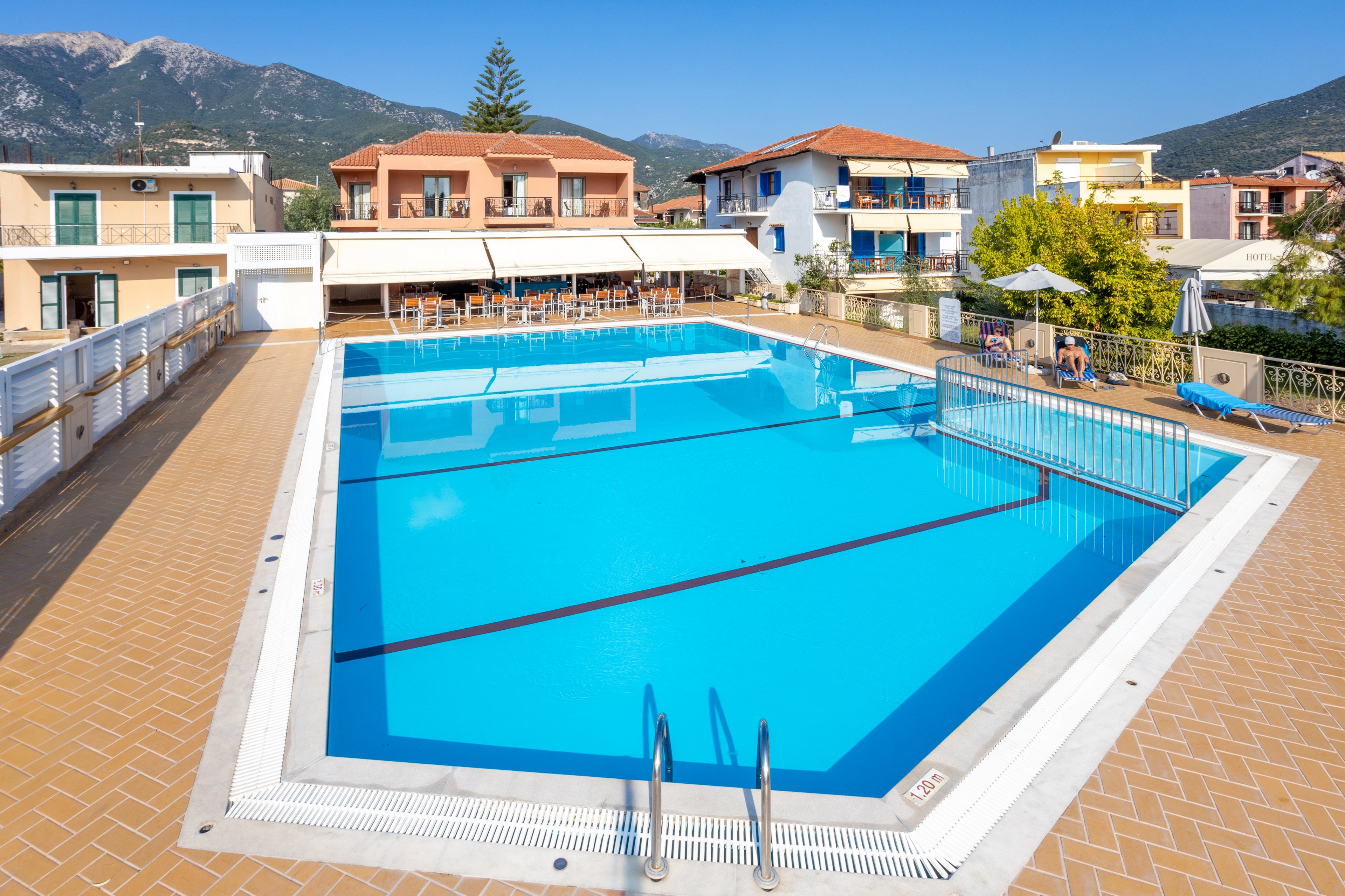 A clear blue swimming pool surrounded by a tiled deck, located in a residential area with colorful buildings. The setting features a sunny day with a mountain range visible in the background. Poolside seating and umbrellas are present, adding to the inviting atmosphere.
