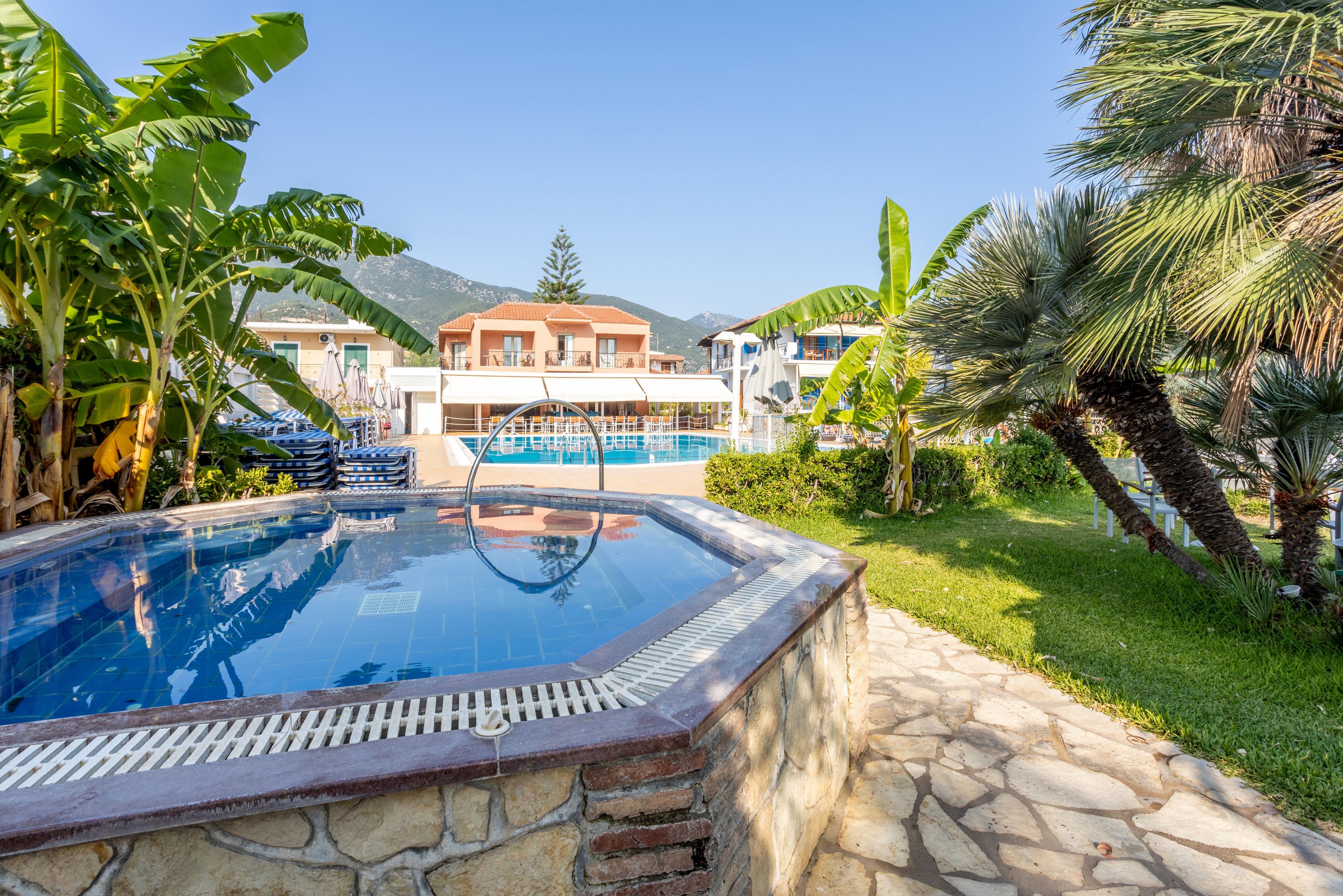 A serene villa featuring a small pool surrounded by lush tropical plants, including banana trees and palms. The setting includes a clear view of distant mountains under a bright blue sky. The villa architecture showcases a Mediterranean style with white and pastel tones, creating a relaxing atmosphere.