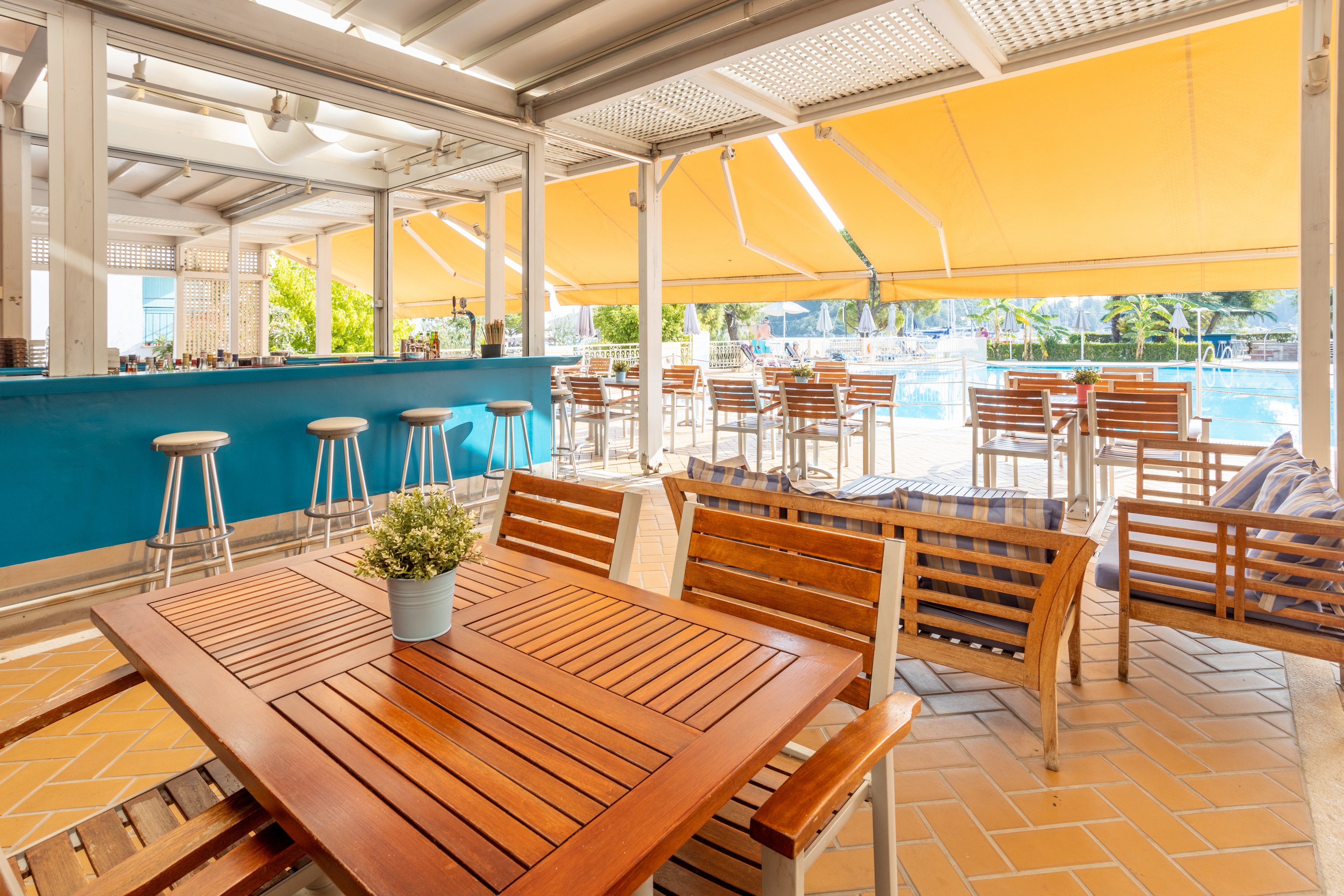 A bright and inviting outdoor café featuring wooden tables and chairs under a yellow canopy. The setting overlooks a sparkling swimming pool, with a teal bar area and metal stools in the background. The space is bathed in natural light, creating a relaxed and airy atmosphere.