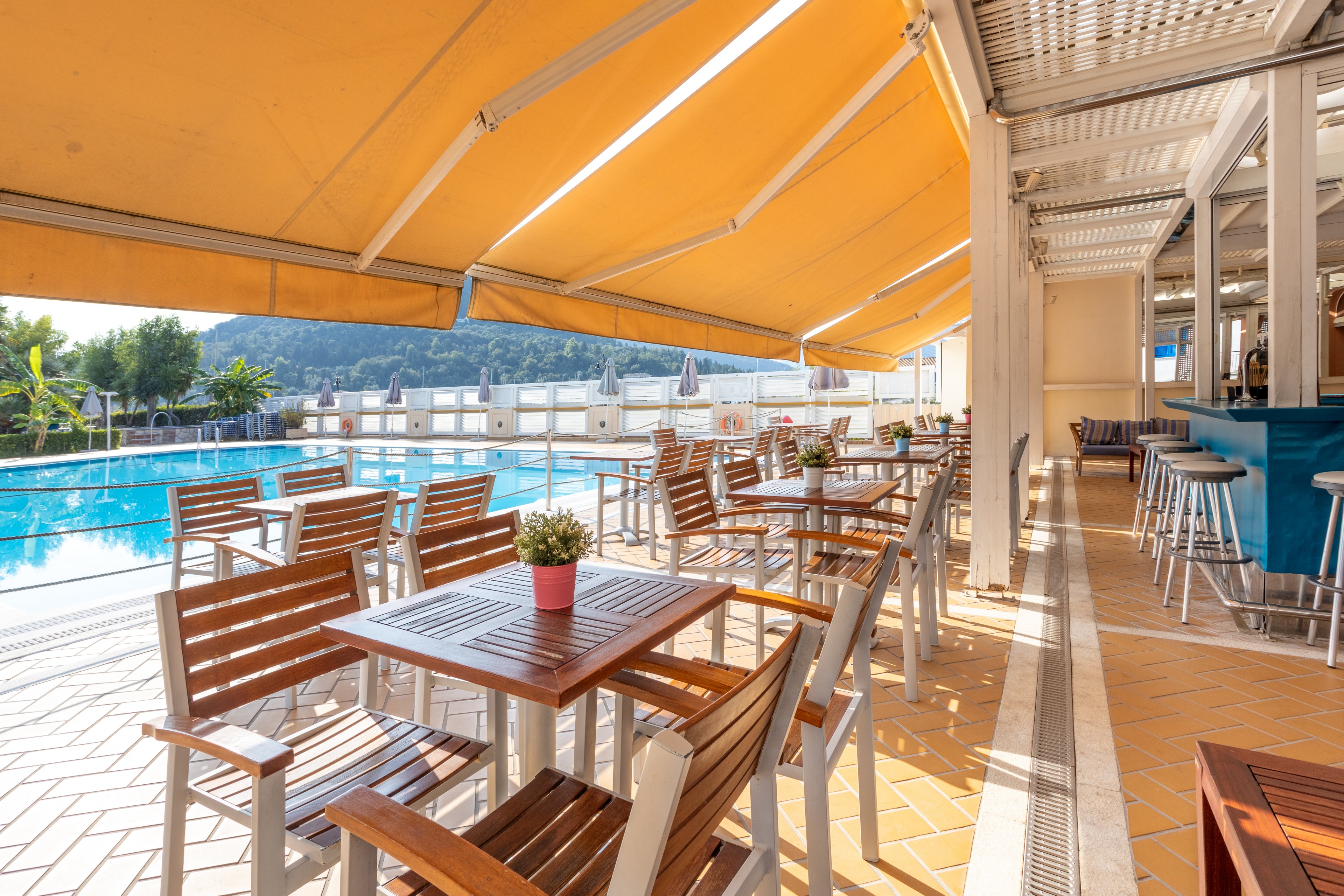 An inviting outdoor seating area featuring wooden tables and chairs under yellow awnings. The setting overlooks a large swimming pool surrounded by greenery and distant hills. Potted plants add a touch of decor, and the area is bathed in natural sunlight.
