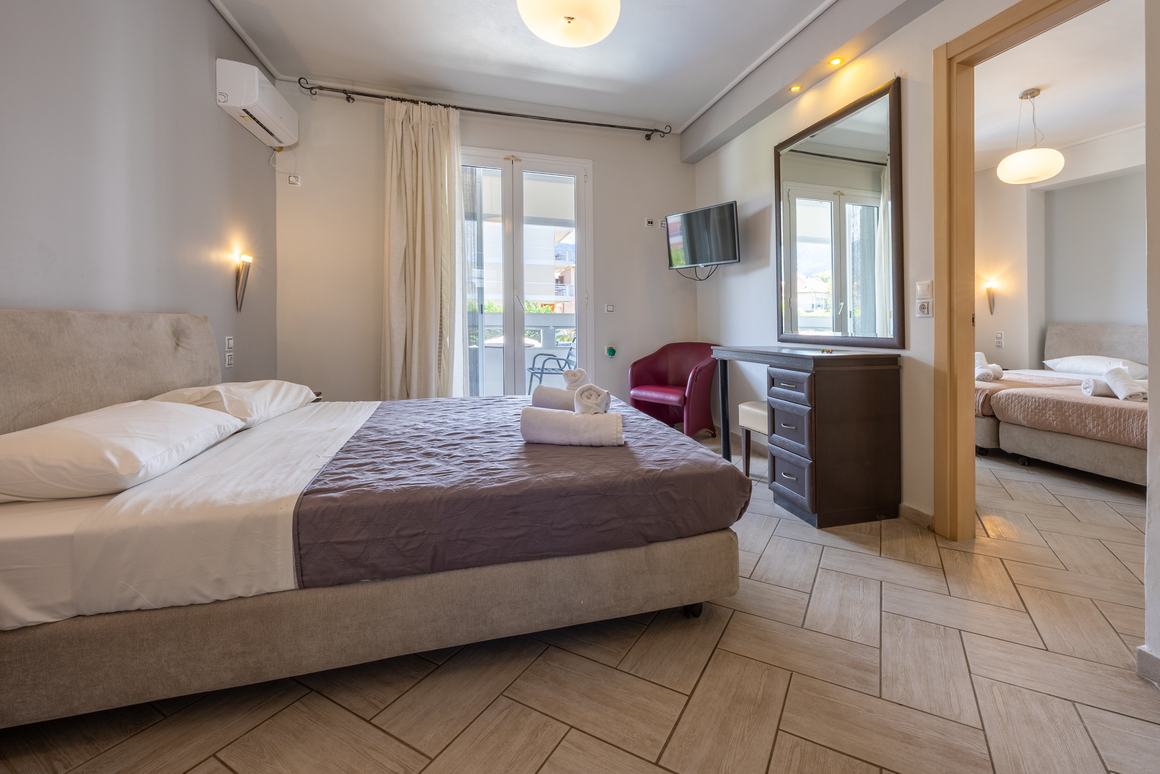 A contemporary hotel room features a neatly made double bed with folded towels placed on top. The space includes a wall-mounted television, a desk with a mirror, and a red armchair near the bed. Large glass doors with light curtains open to a balcony, allowing natural light to brighten the neutral-toned interior. Through an open doorway, a second bedroom with another bed and soft lighting is visible, suggesting a connected suite layout.